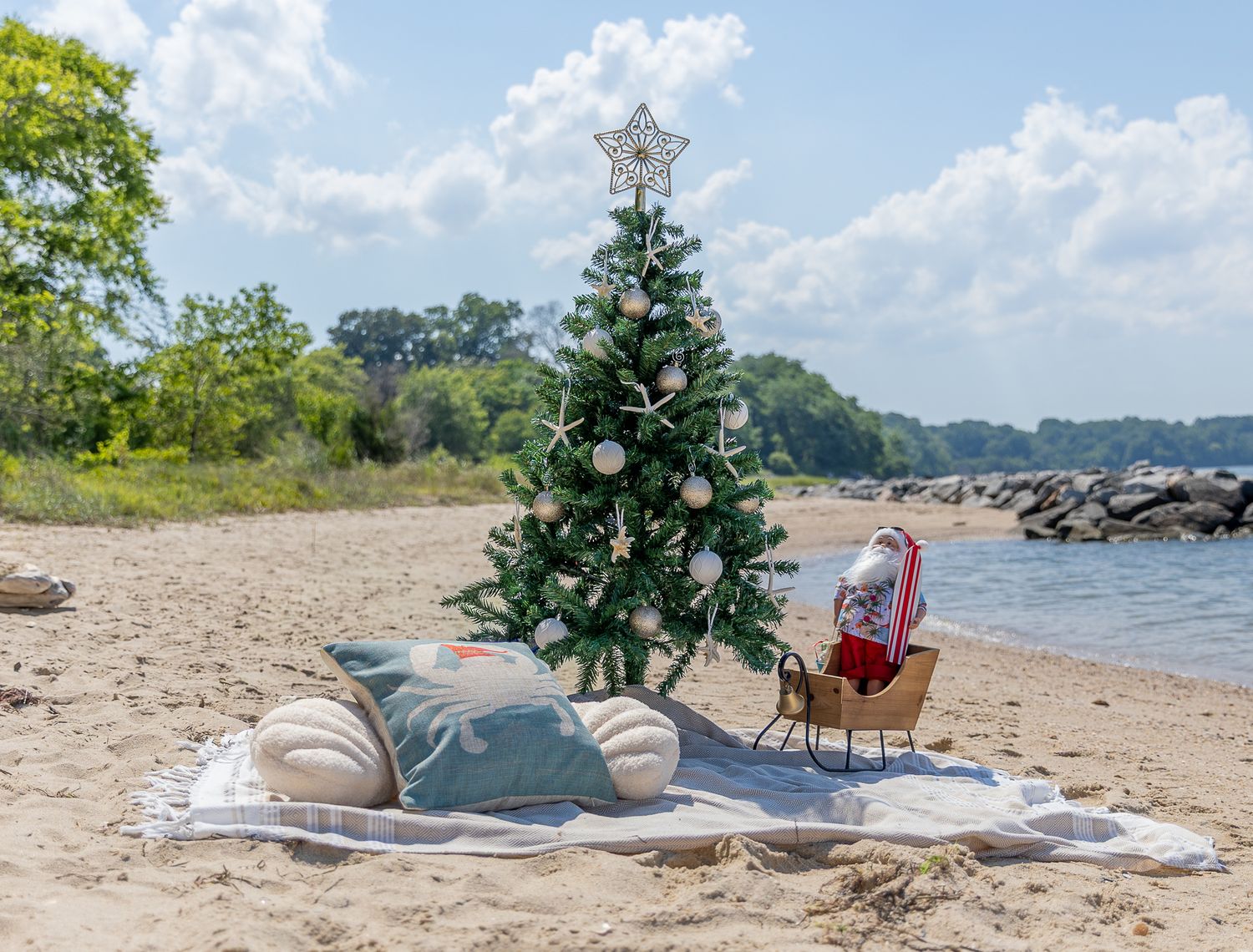 A decorated Christmas tree with a Santa sleigh figurine on a sandy beach blanket by the water.