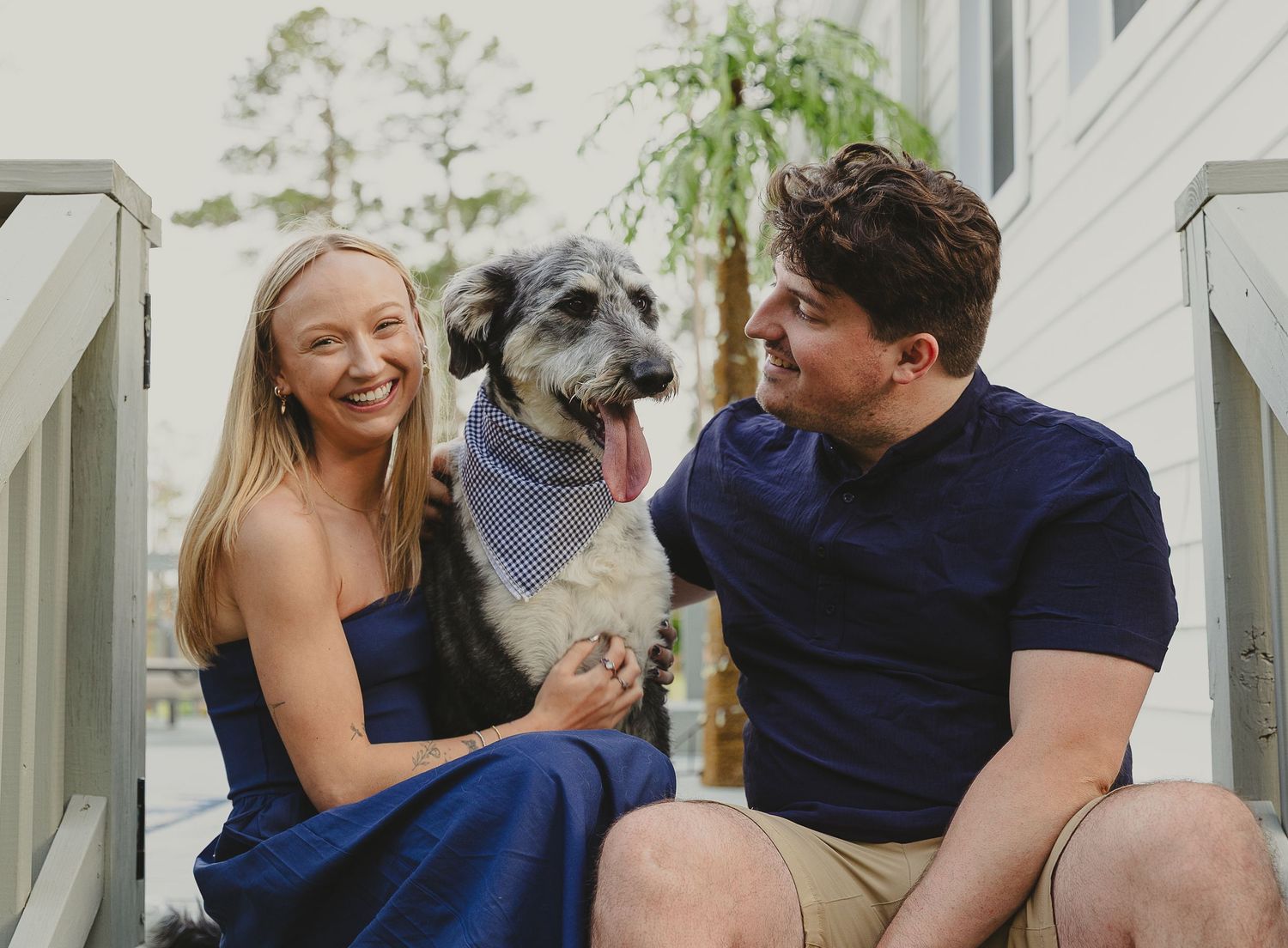 A smiling couple sits on porch steps with a fluffy dog wearing a bandana between them.