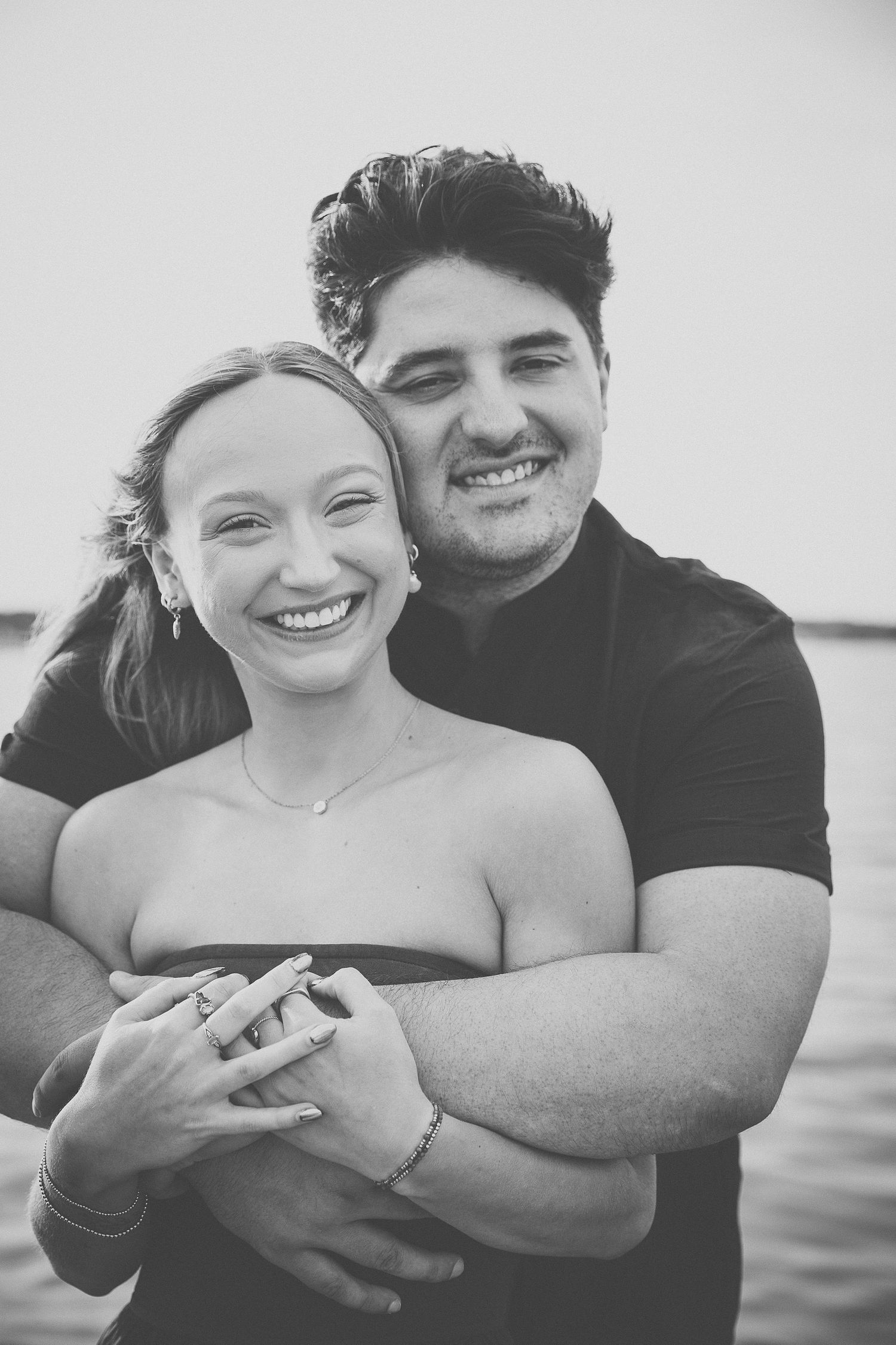 Black and white photo of smiling couple embracing near the water, woman wearing engagement ring.