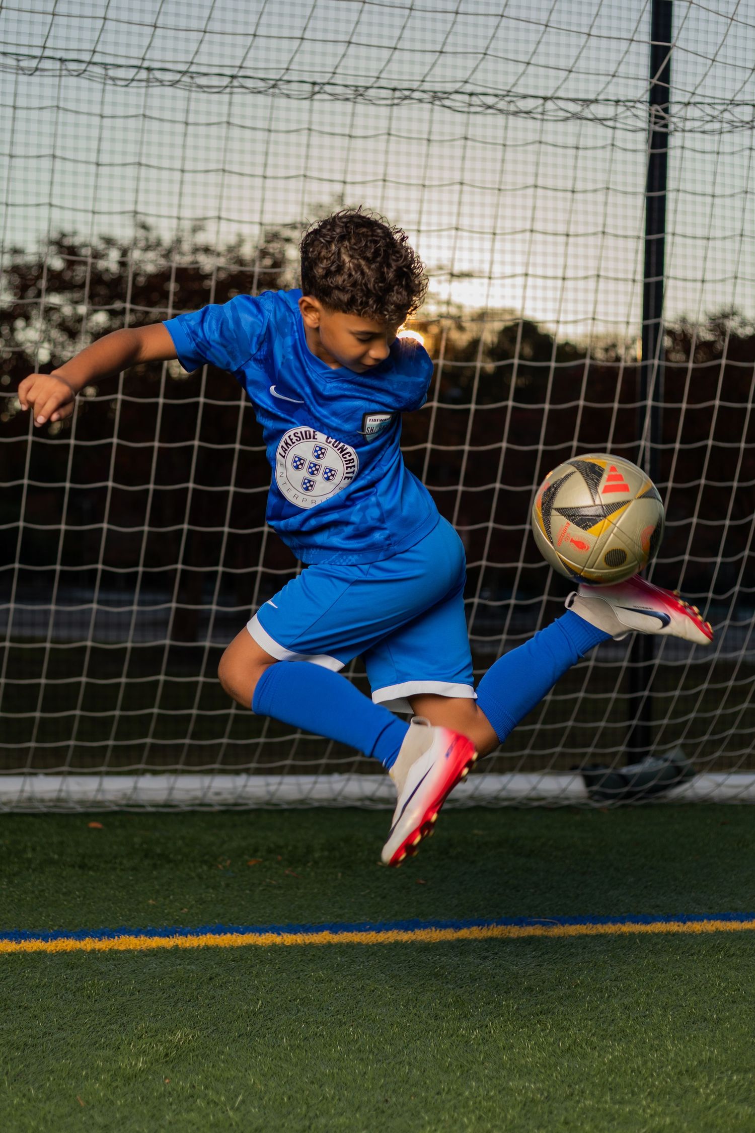 Young soccer player in blue uniform leaping mid-air to kick ball in front of goal net on green field.