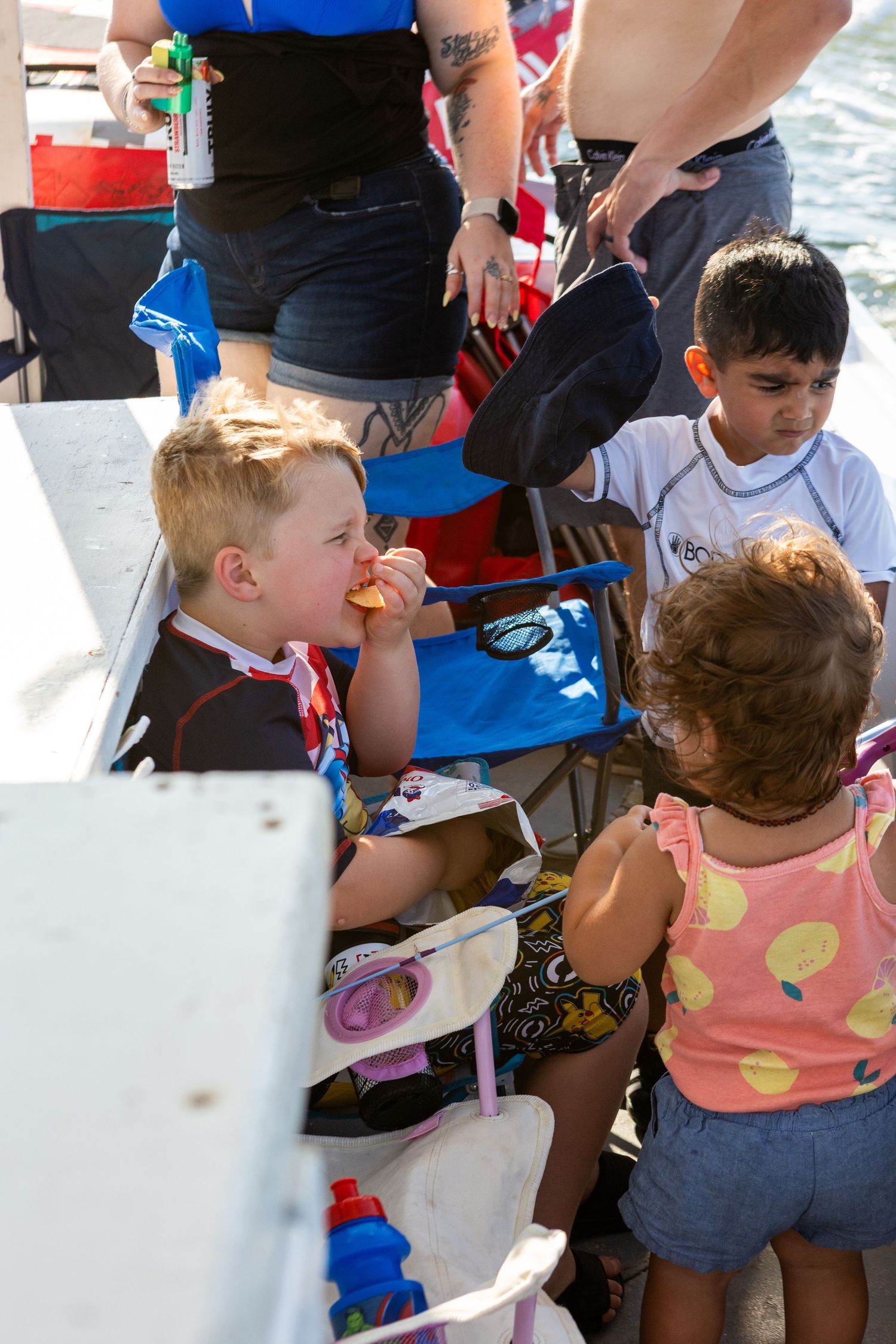 Children eating snacks outdoors at a sunny waterside event, with adults nearby and colorful chairs in the background.