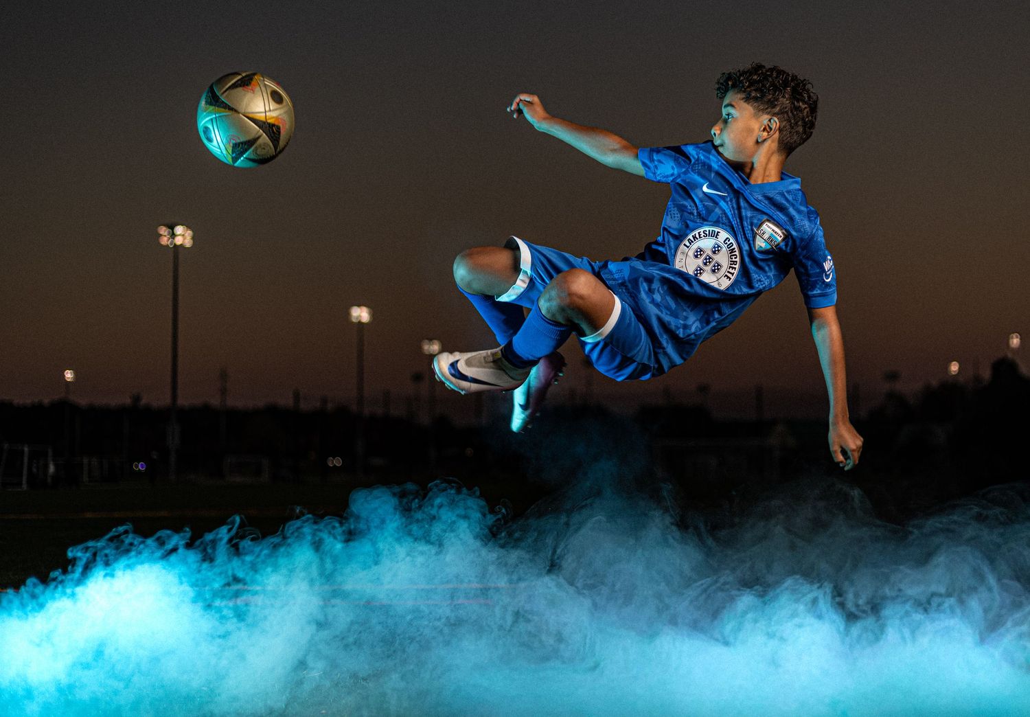 Young soccer player in blue jersey performs dramatic bicycle kick surrounded by teal smoke under night sky.