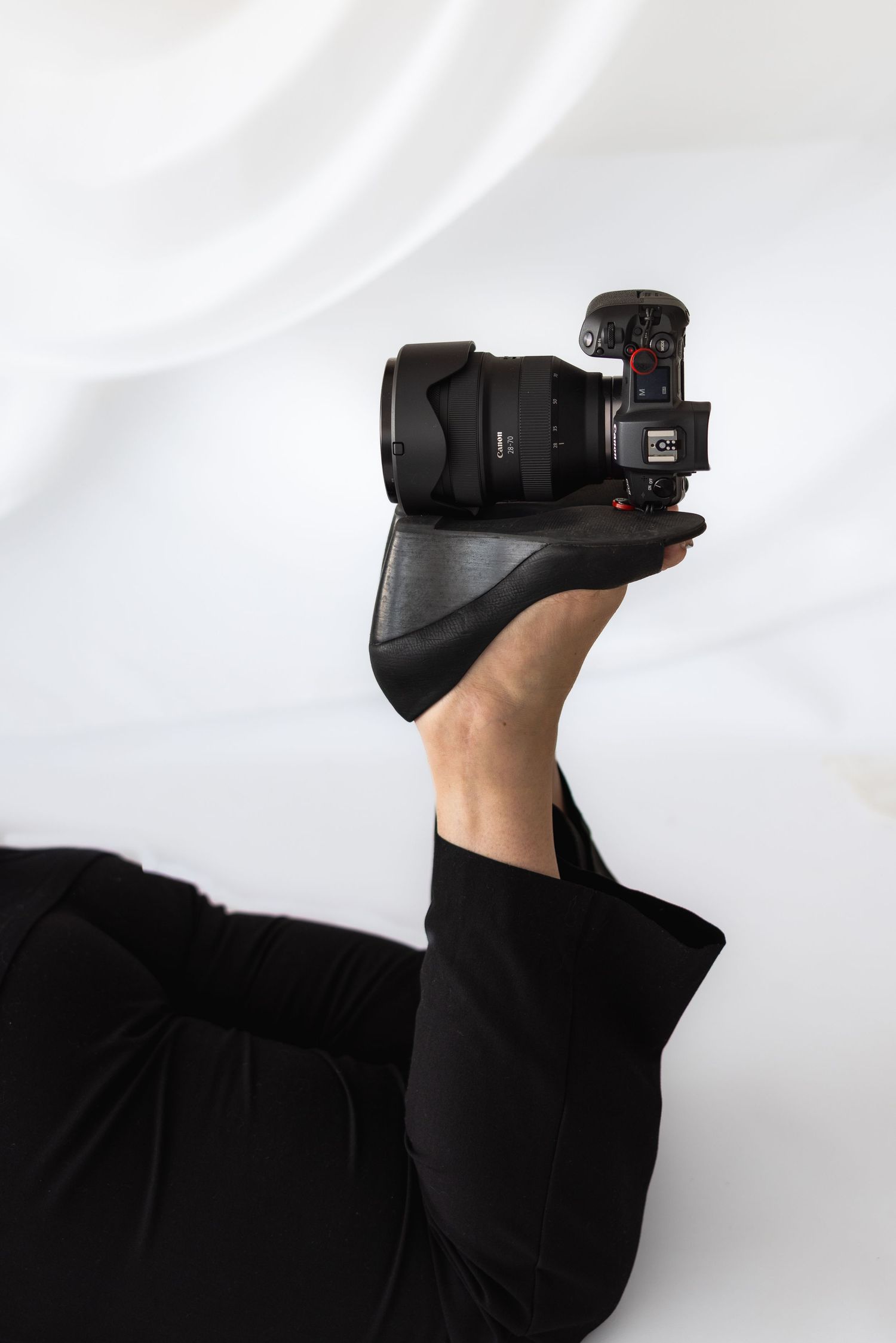 Person in black outfit holding a professional DSLR camera against white fabric background.