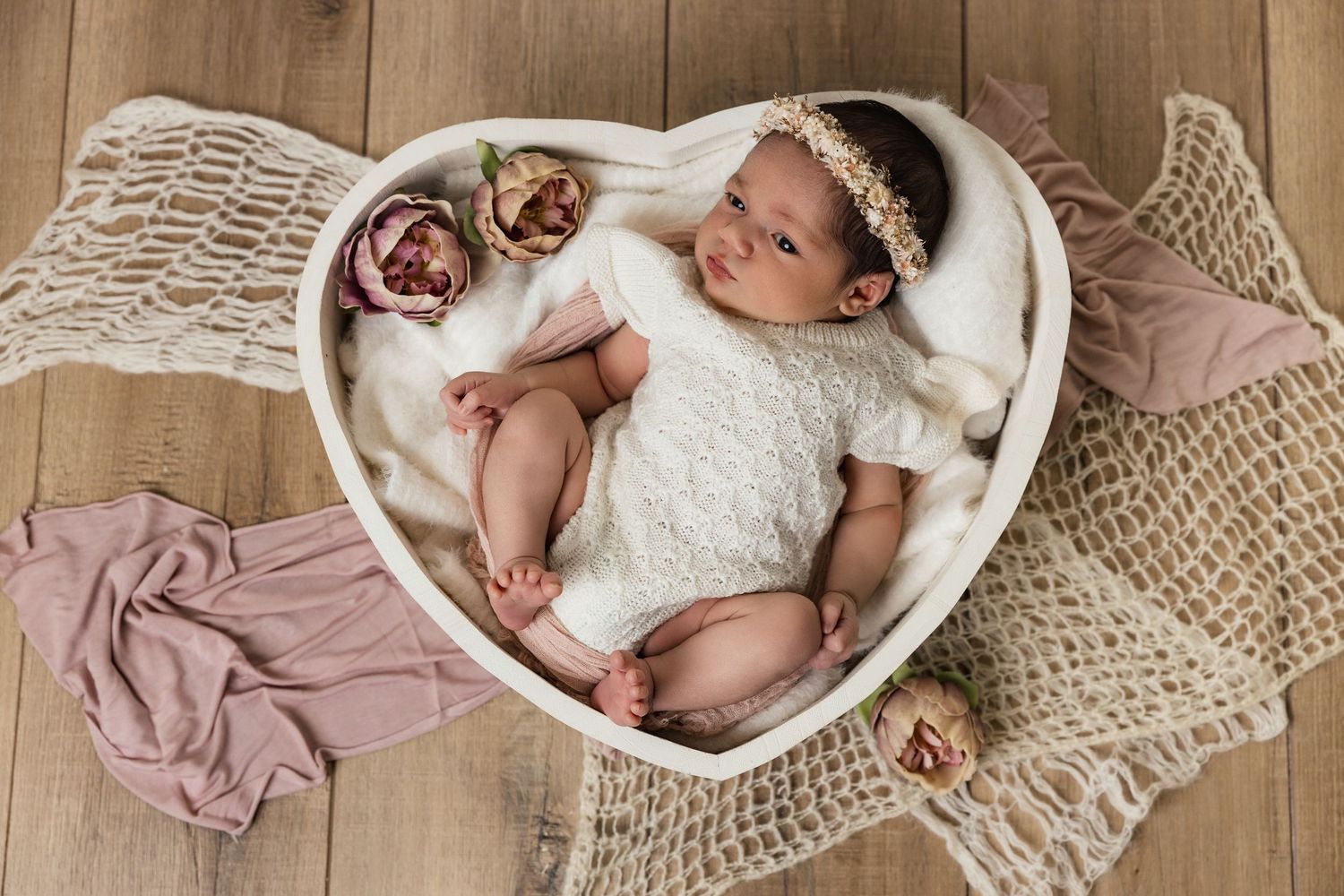 Newborn baby in white lace outfit sleeping in a heart-shaped bowl with floral accents.