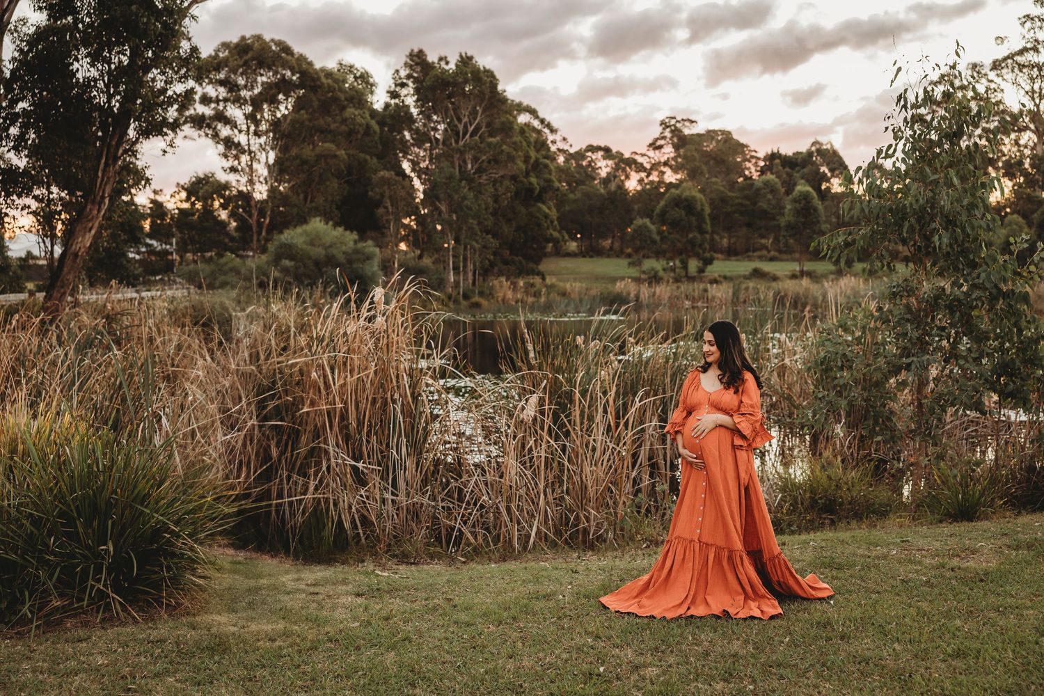 Pregnant woman in flowing orange gown posing in lush green garden at golden hour sunset.