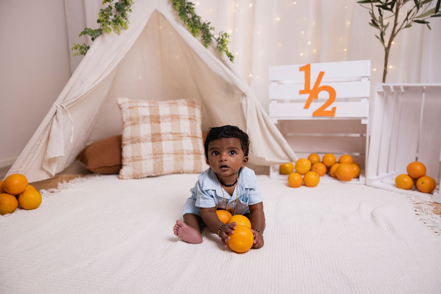 Baby boy sitting near white teepee tent surrounded by oranges celebrating half birthday milestone.
