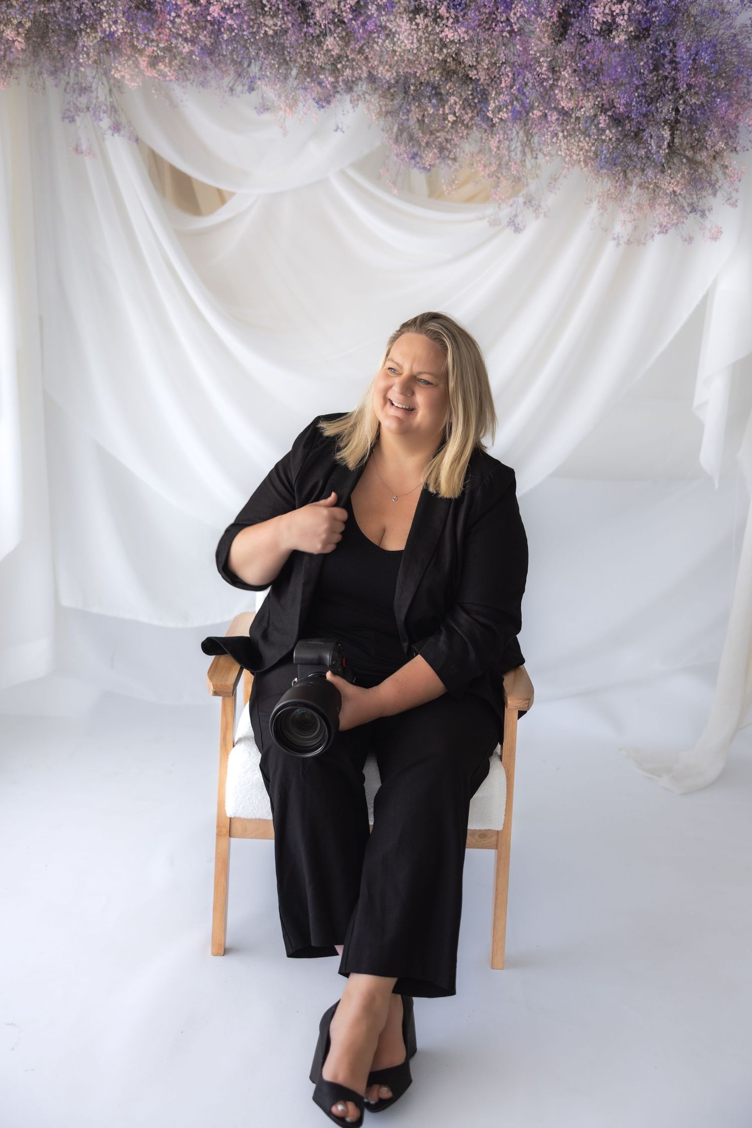 Smiling female photographer in black blazer seated in chair holding professional camera in studio.