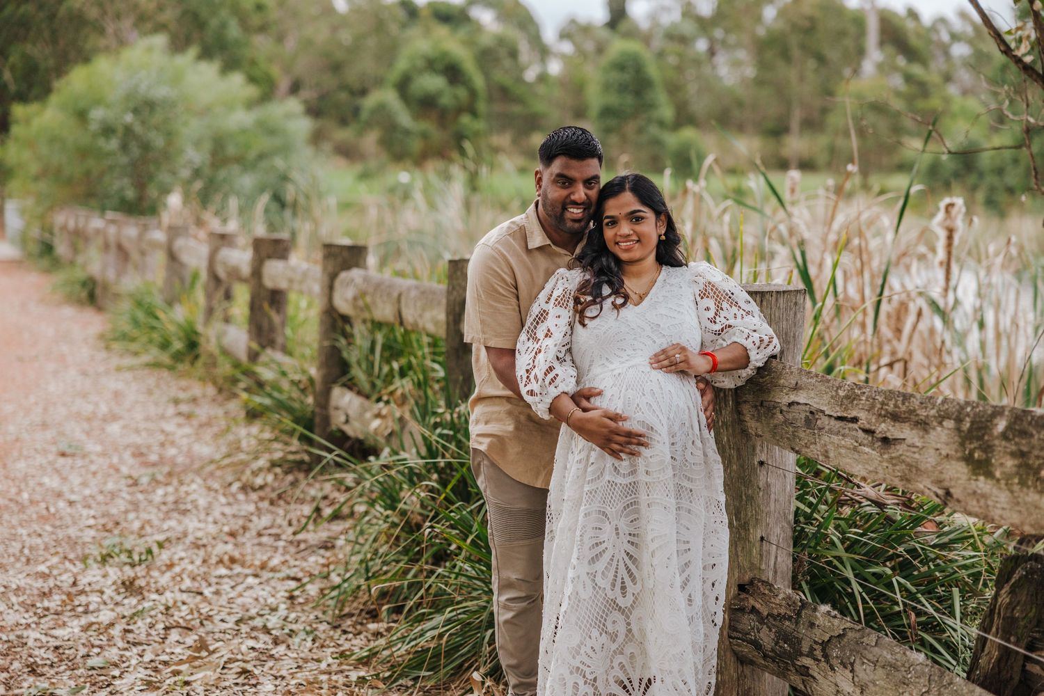 Expecting couple posing outdoors near wooden fence and pond for maternity photography session.