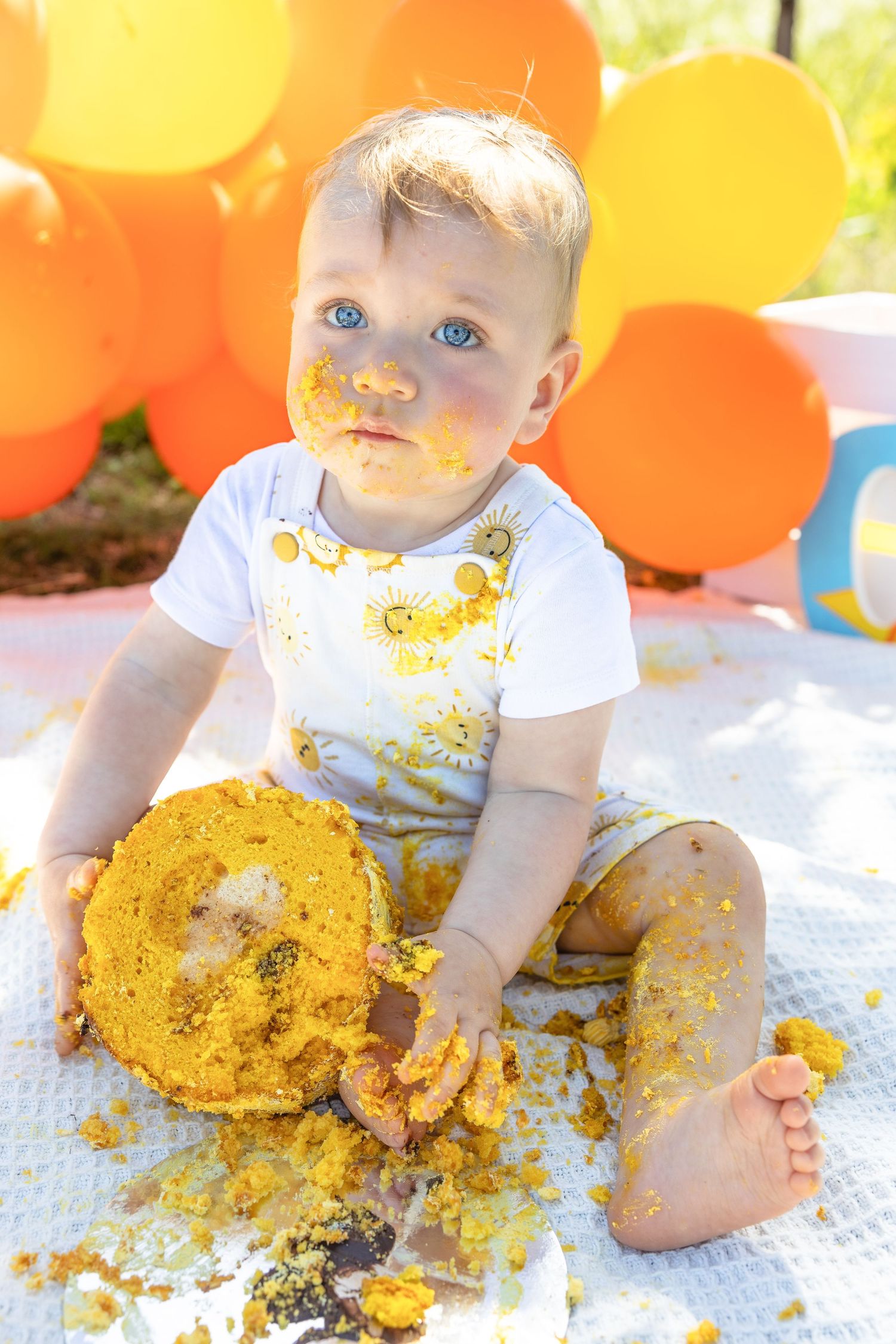 Baby covered in yellow cake celebrating first birthday with orange and yellow balloons outdoors.