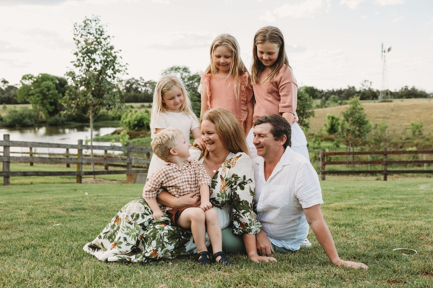 Joyful family of six sitting together on green lawn near fence laughing and embracing.