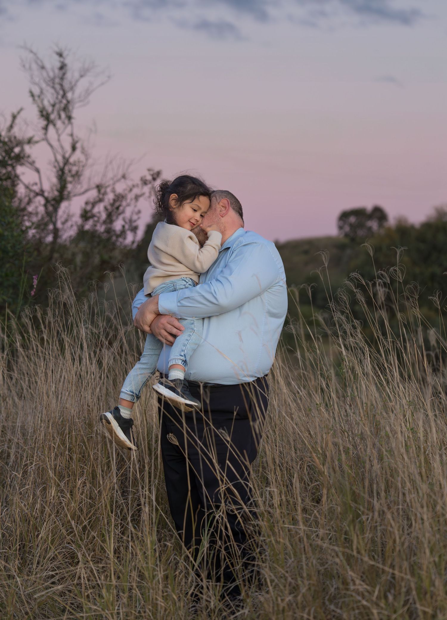 Father holding young daughter in tall grass field at sunset with pink sky background.