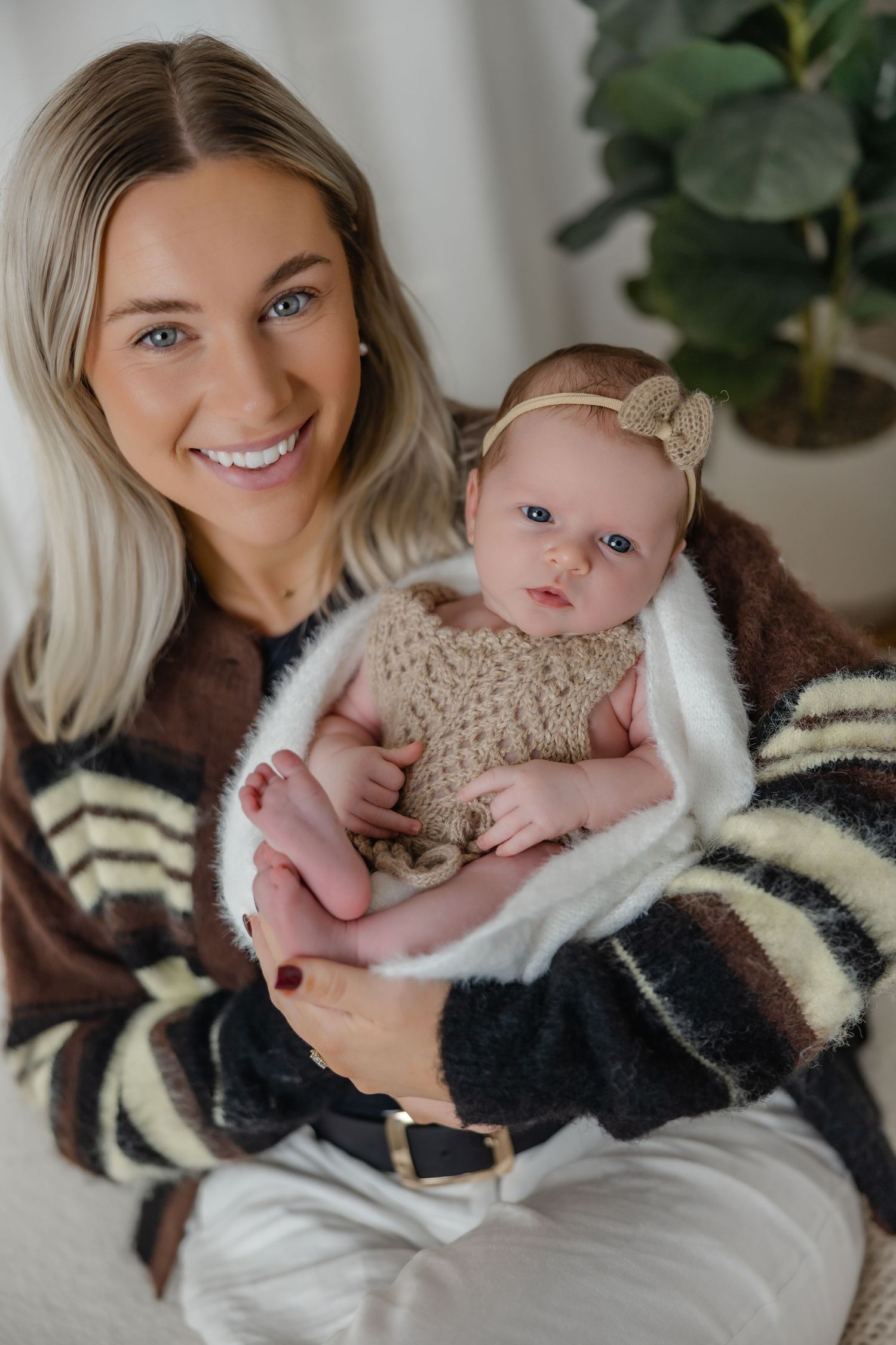 Smiling blonde mother holding newborn baby in knitted outfit indoors.
