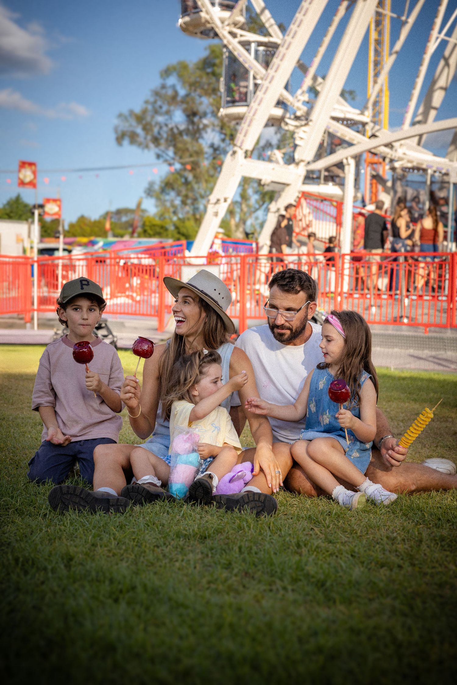 Happy family sitting on grass enjoying candy apples near a ferris wheel at a fair.