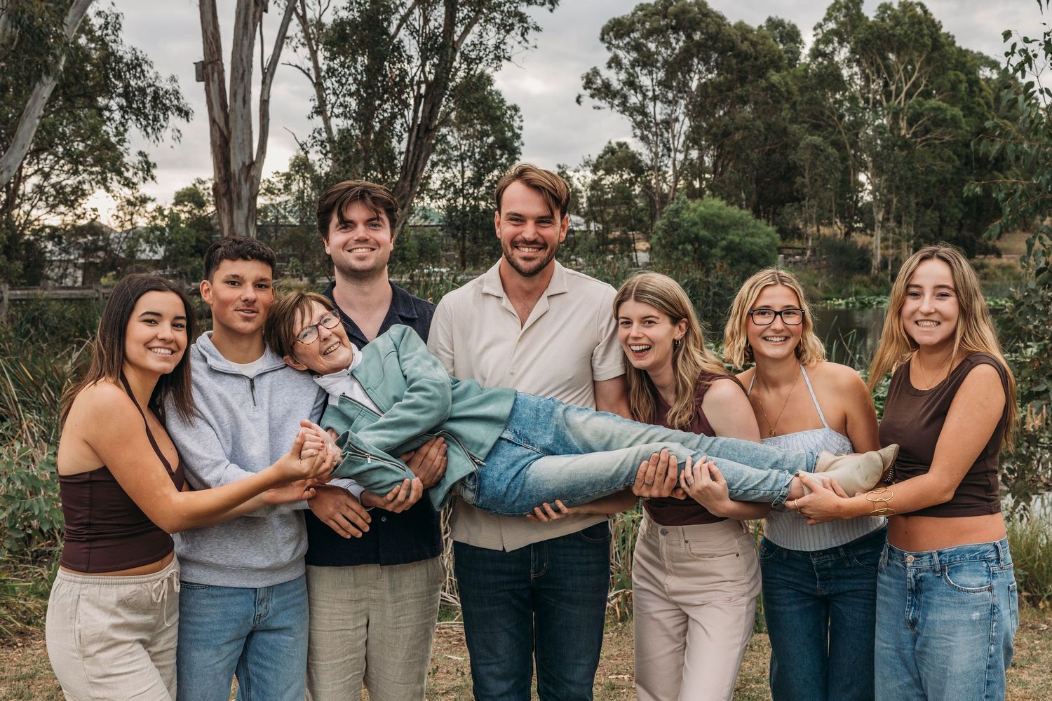 Group of seven young adults smiling together outdoors in a park setting with trees in the background.