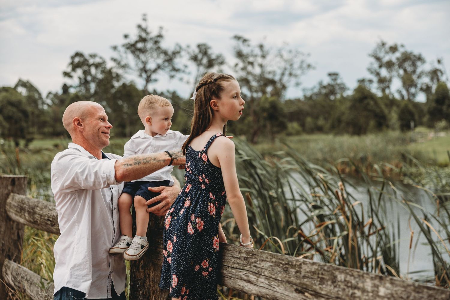 Father holding toddler son while pregnant mother stands by rustic wooden fence near pond.
