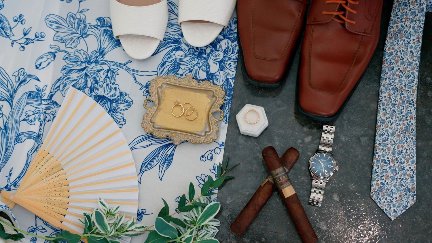 Wedding accessories including shoes, fan, tie, and watch arranged on blue floral fabric.