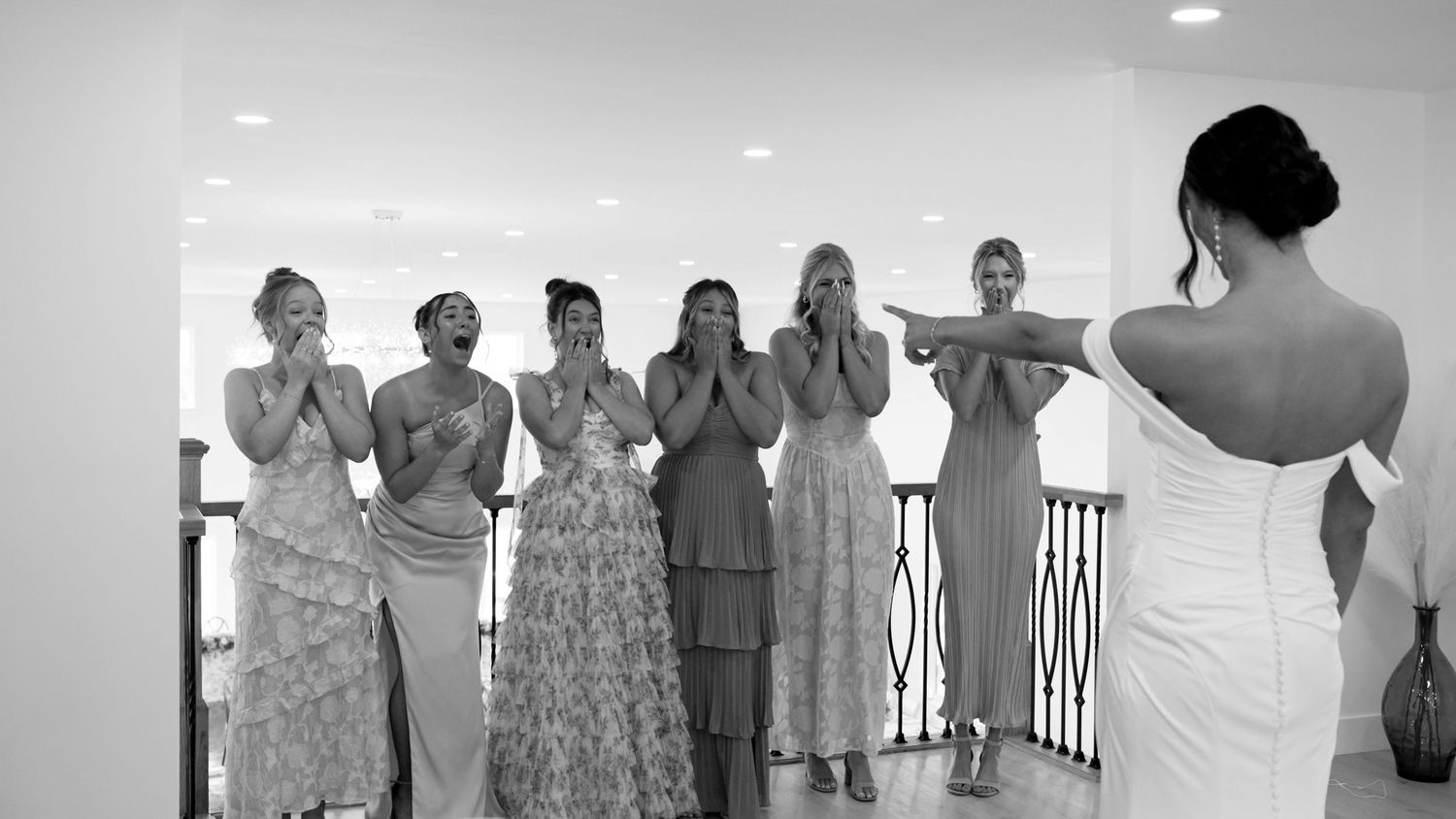 Bridesmaids in floral dresses and formal attire react with joy and surprise during a wedding photoshoot.