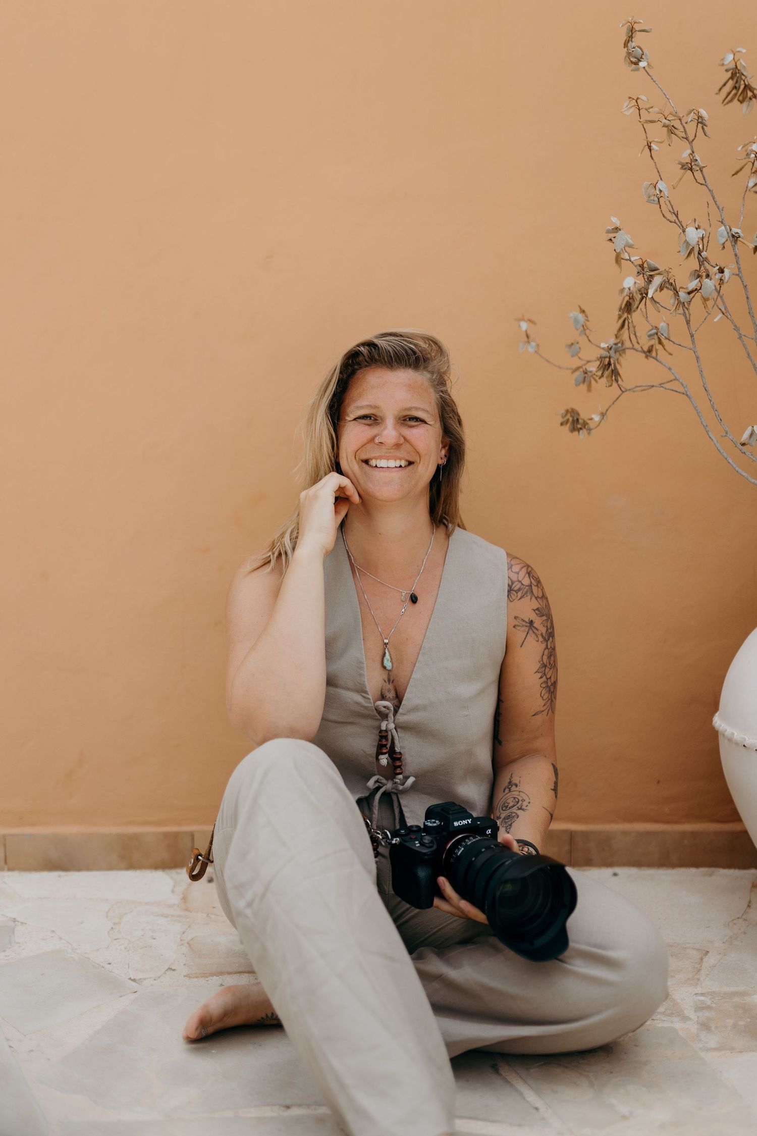 Wedding photographer with camera sits casually on floor in beige studio setting.