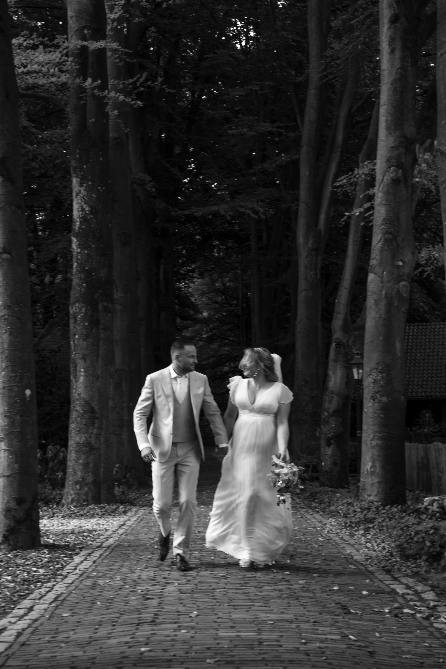 A wedding couple walks along a tree-lined brick path in black and white.