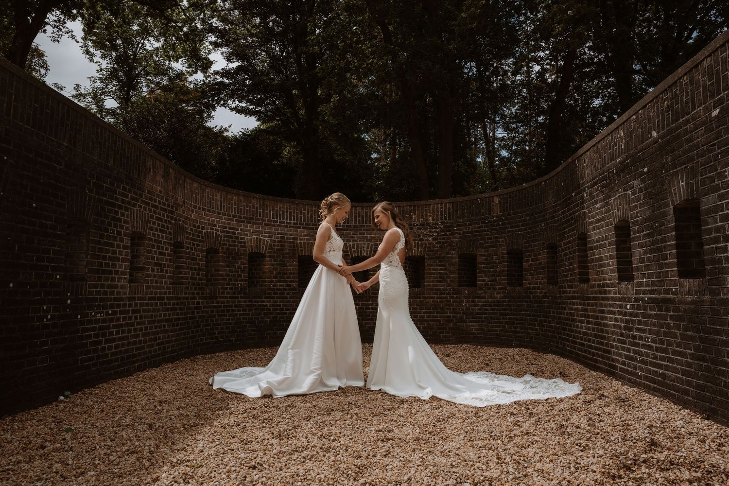 Two wedding dresses with long trains in brick-walled courtyard setting.