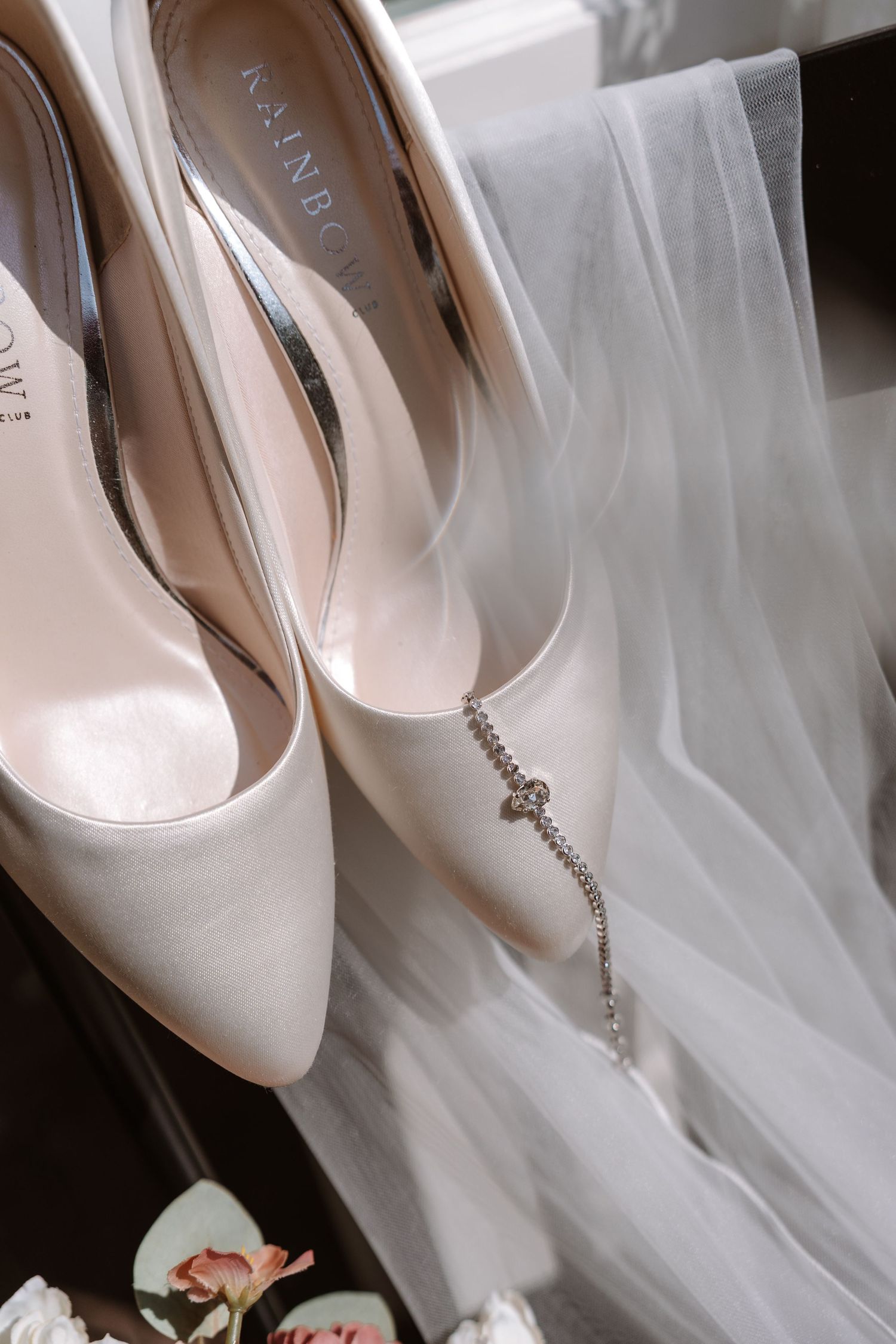 Elegant ivory satin bridal heels with crystal anklet bracelet beside a white wedding veil.