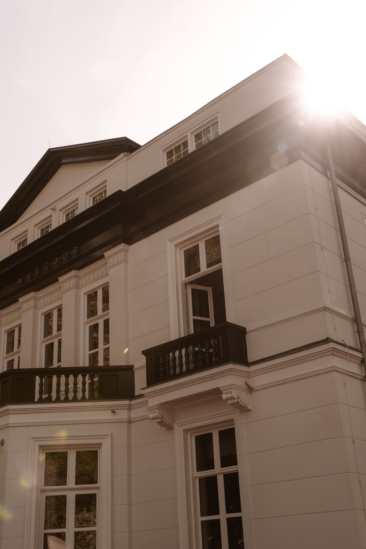 Elegant white classical building with balcony and dark trim, captured from below with sun flare in the background.