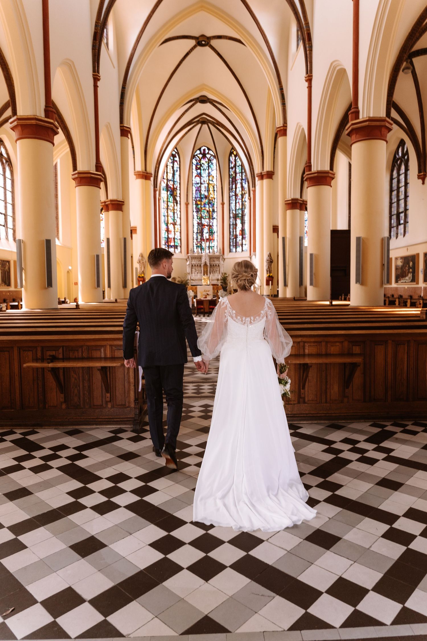 Bride and groom walking hand in hand down a Gothic church aisle with checkered floors and stained glass windows.