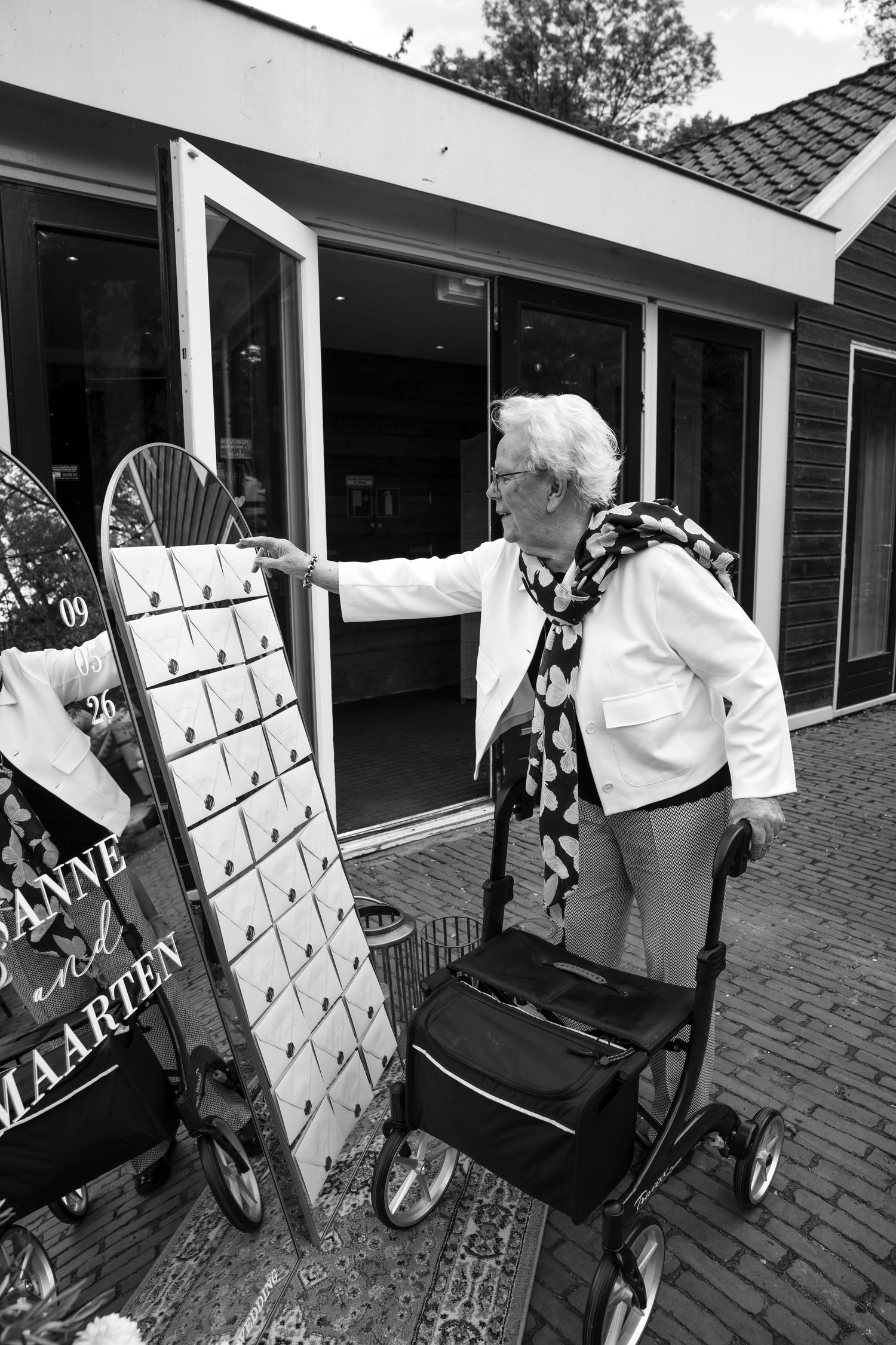 Elderly woman with walker opening door next to decorative sign board outside modern building, black and white photo.