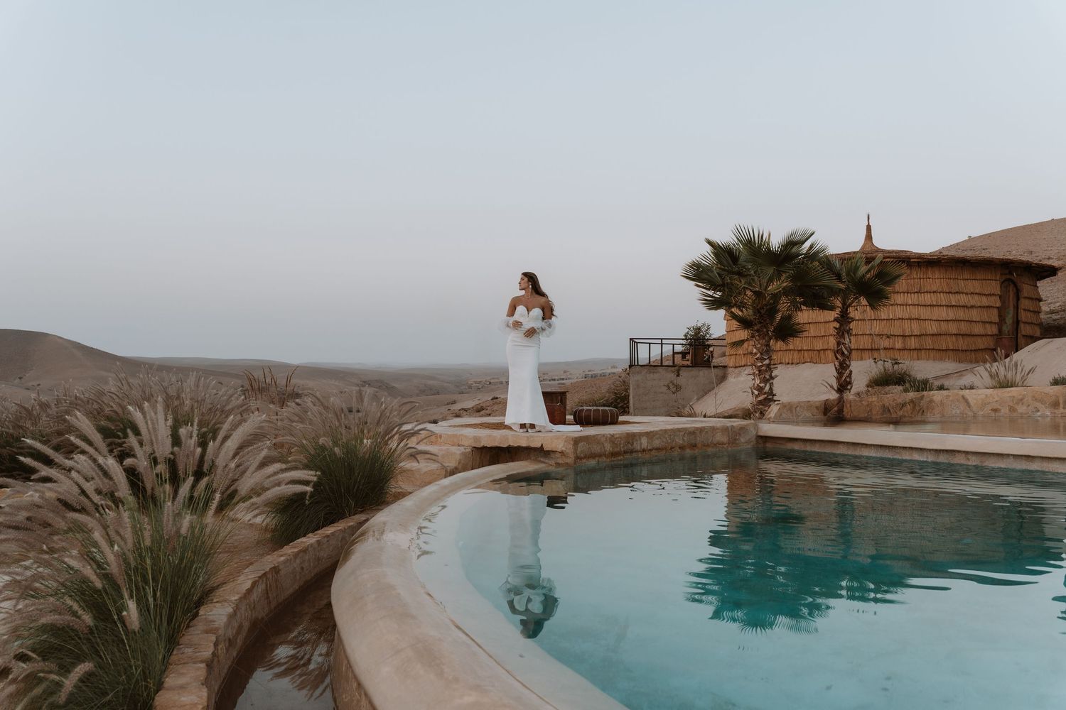 Desert landscape with an infinity pool overlooking mountains and palm trees at sunset.