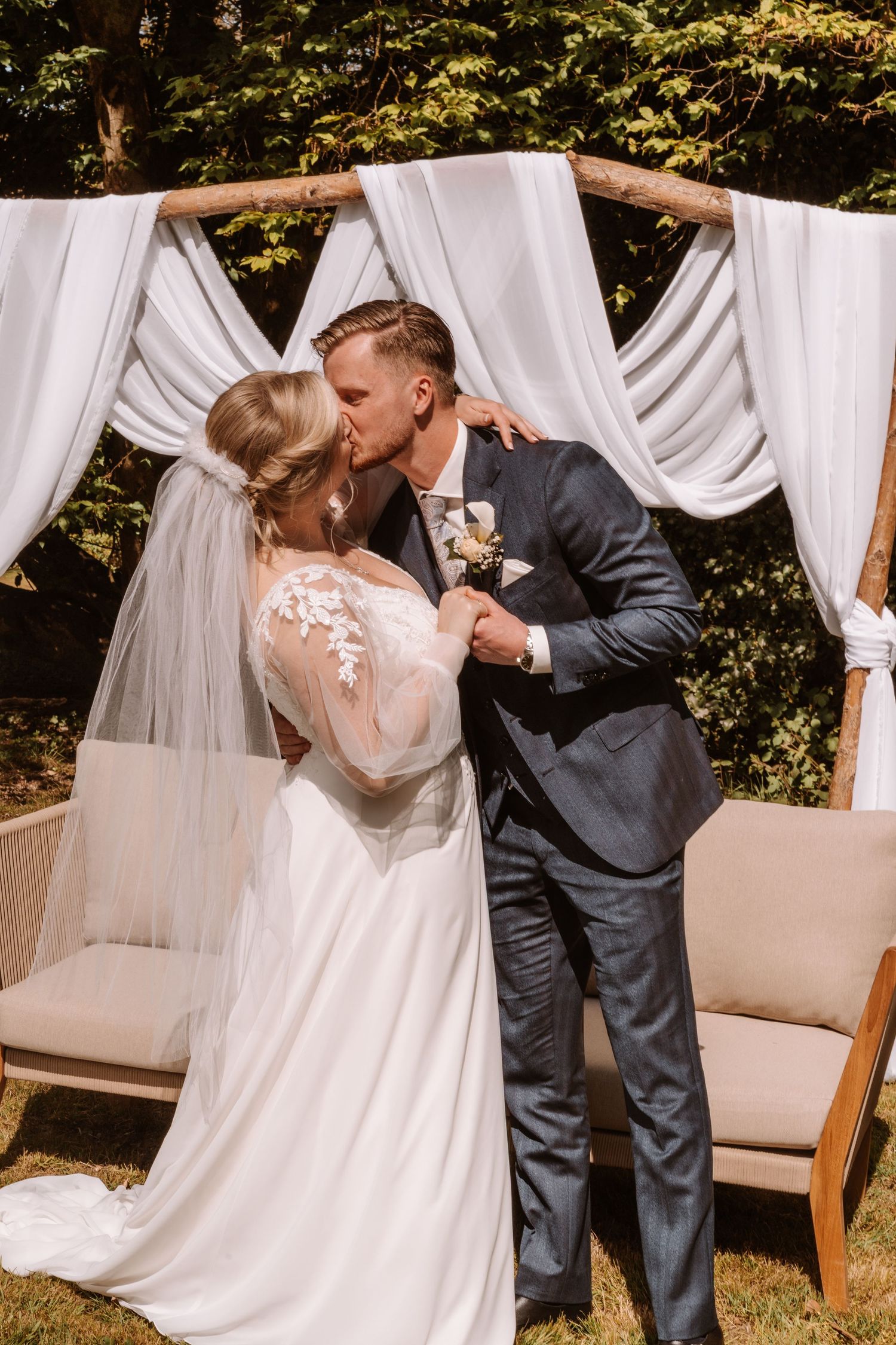 Bride and groom share first kiss at outdoor wedding ceremony beneath white draped arch surrounded by greenery.