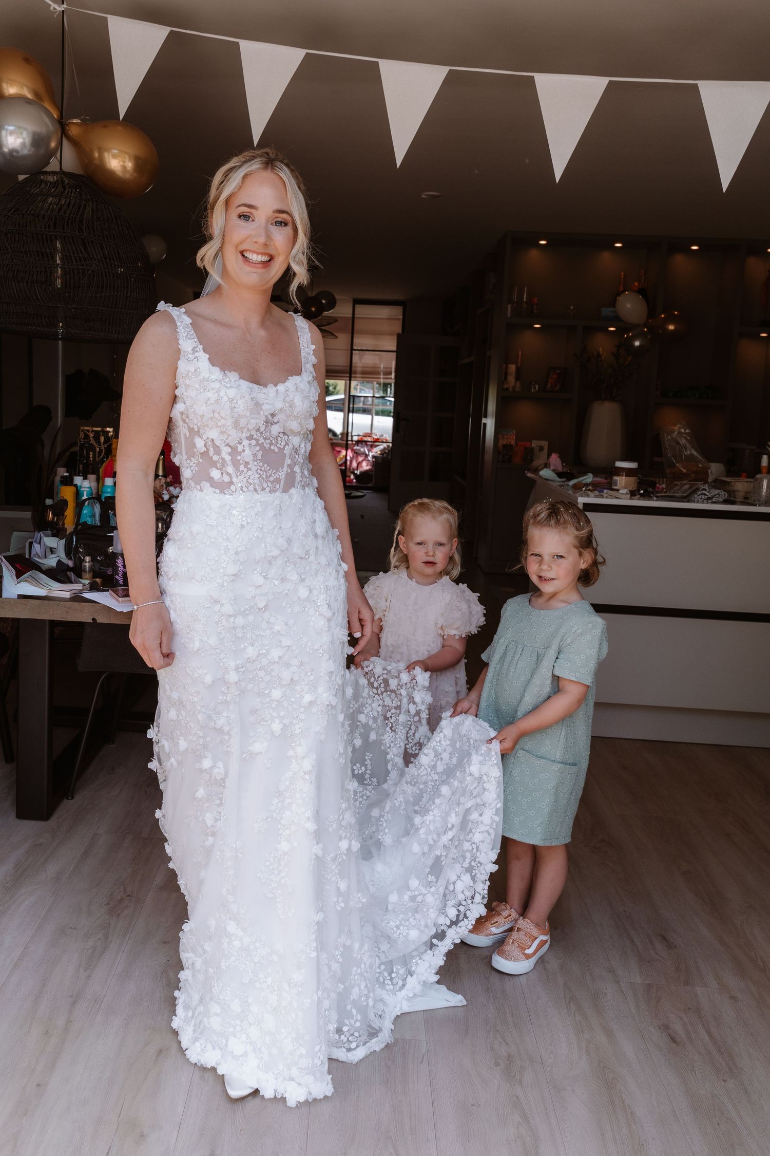 Bride in white floral wedding gown smiles as two young flower girls hold her train indoors.