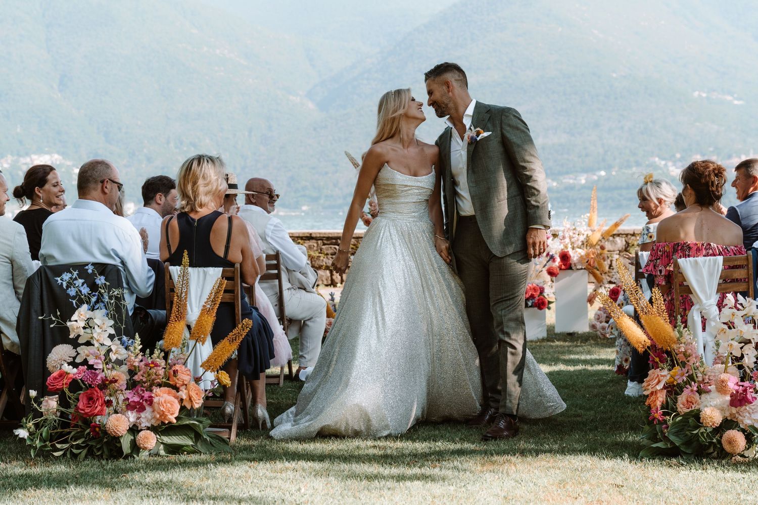 Outdoor wedding ceremony with floral arrangements and guests seated on lawn against scenic mountain backdrop.