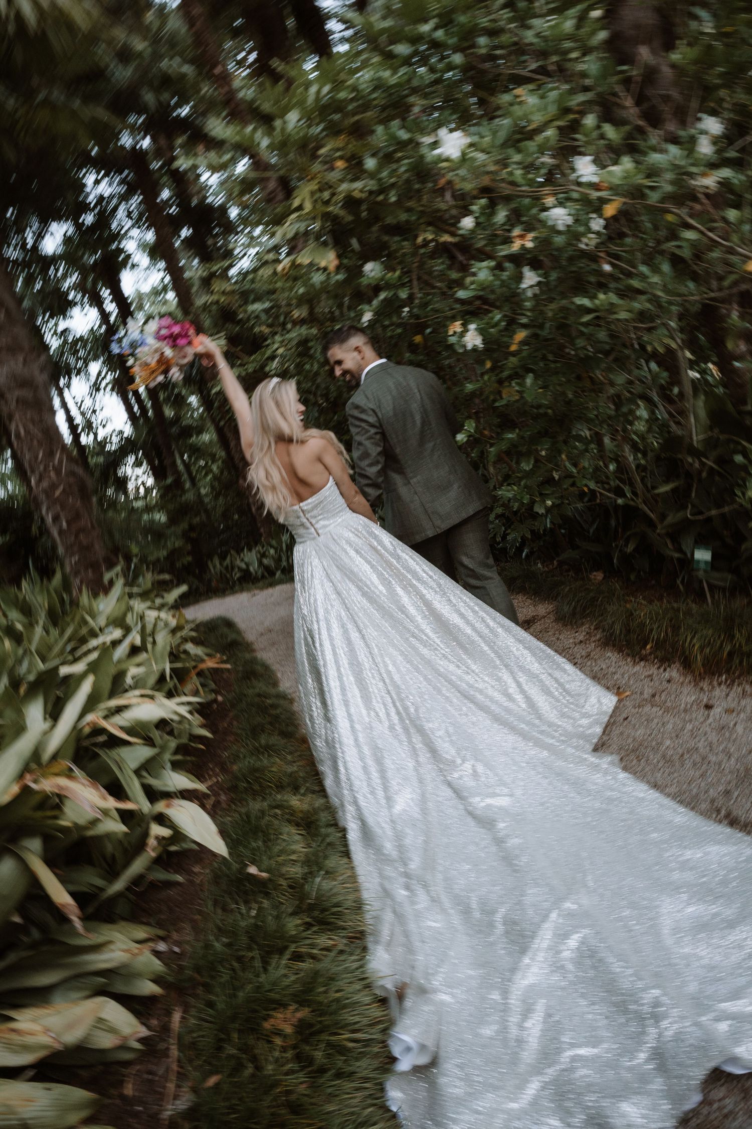 A couple in formal wedding attire embraces in a lush garden setting with a flowing white wedding dress train.