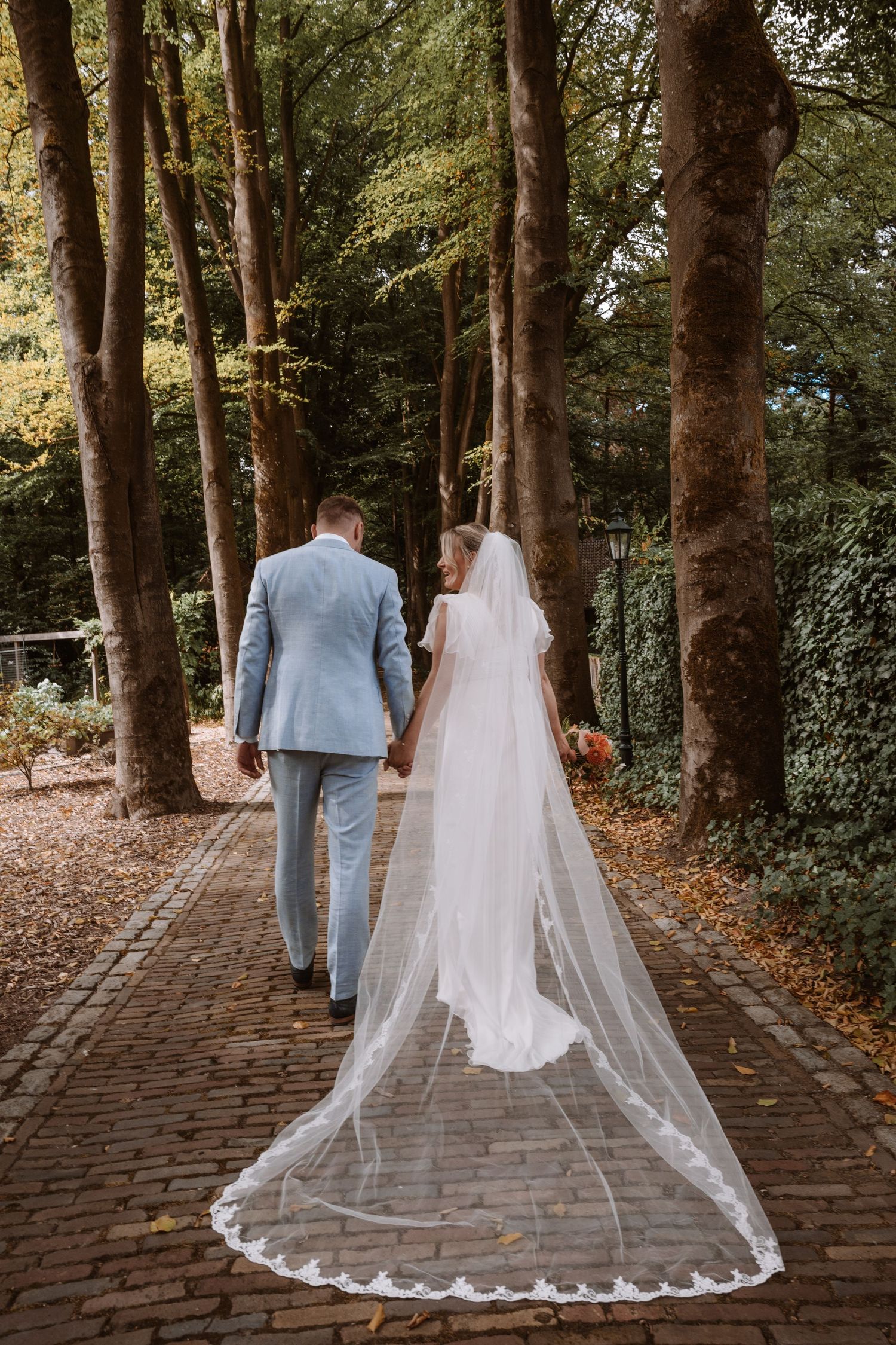 A couple in wedding attire walks down a tree-lined brick path with a cathedral veil trailing behind.