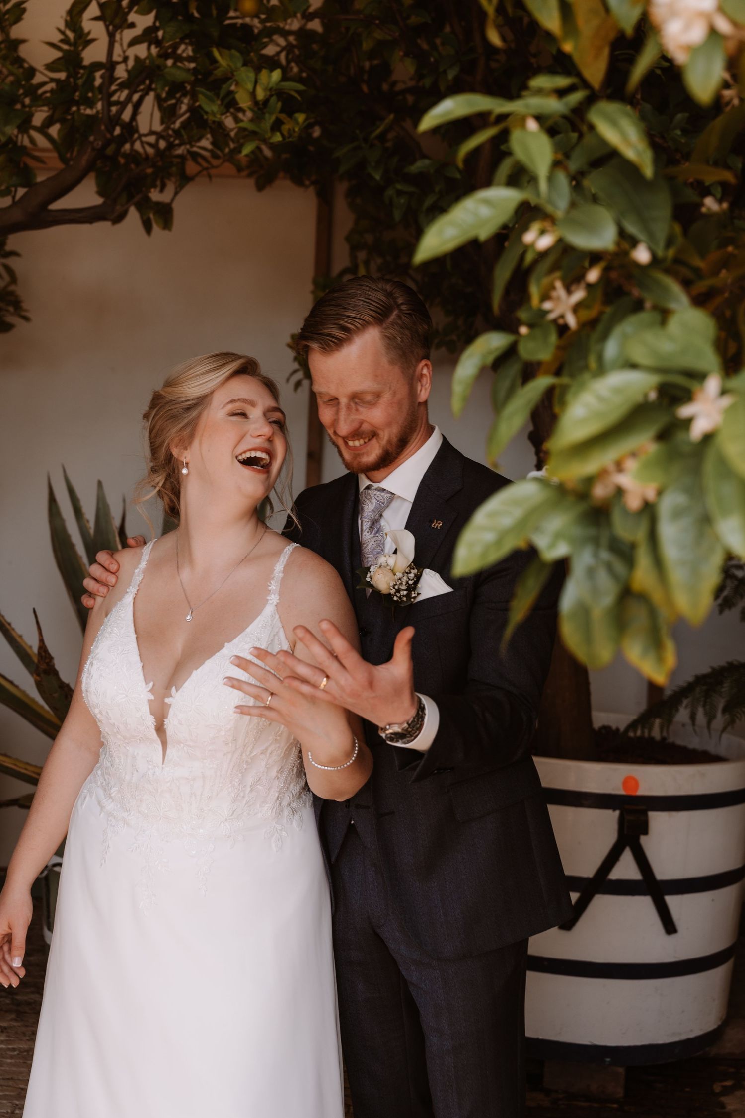 A laughing bride in a white lace gown and groom in a navy suit share a joyful moment surrounded by lush greenery.