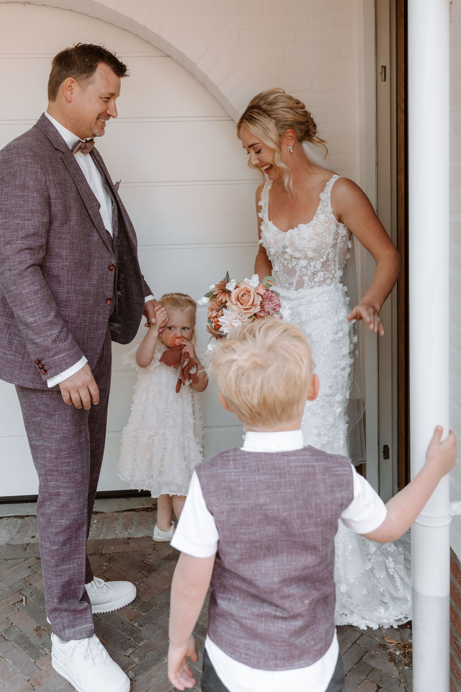 Bride in white lace gown and groom in gray suit greet two young children at church entrance on wedding day.