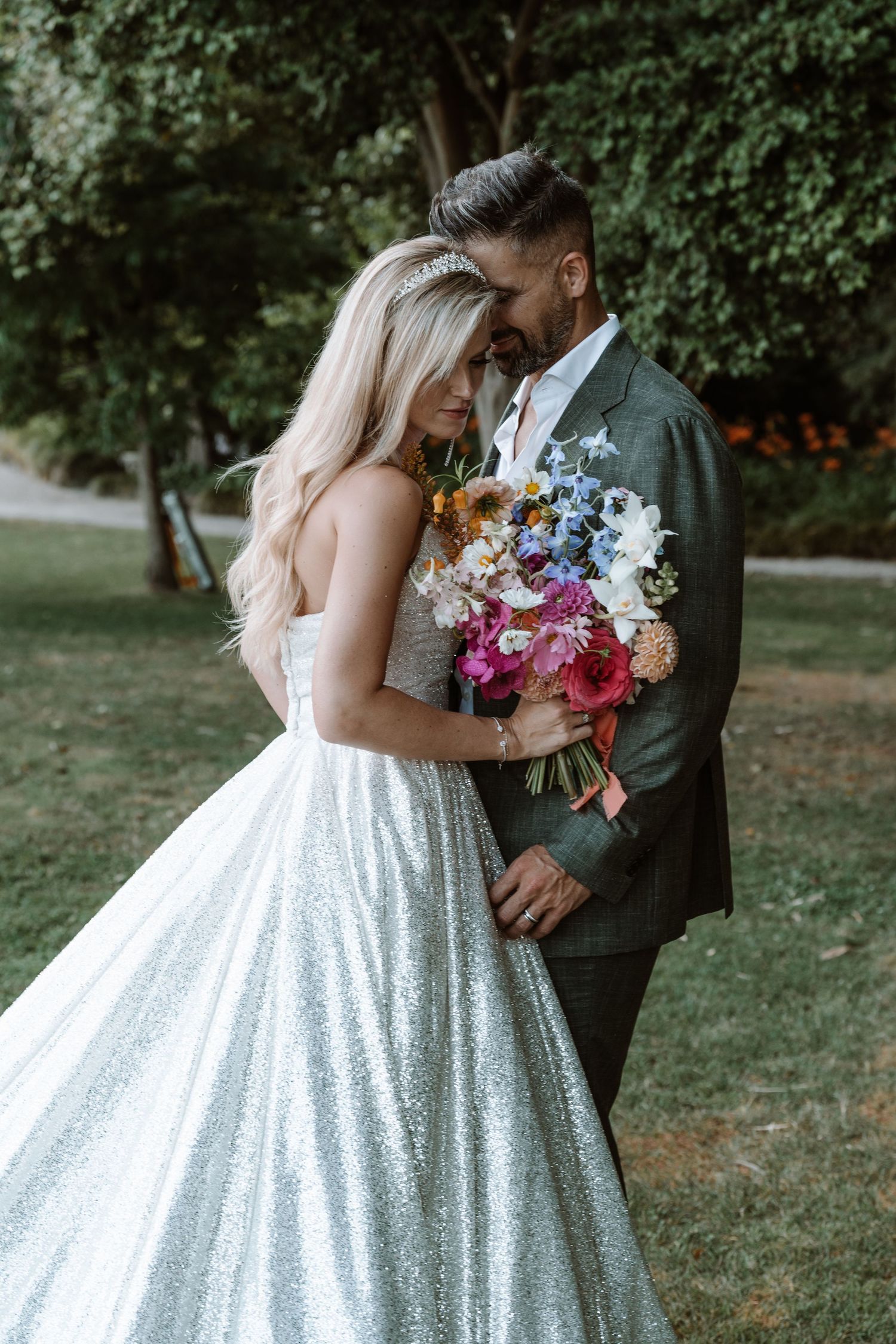 A romantic wedding couple embraces outdoors surrounded by greenery while holding a vibrant floral bouquet.