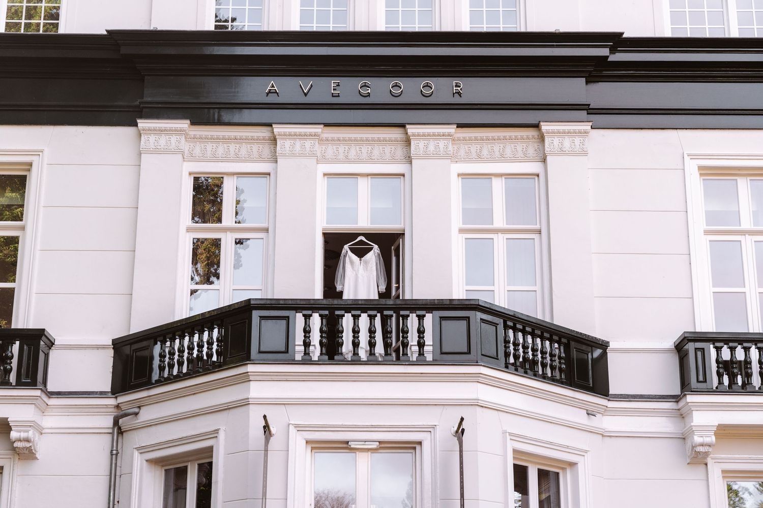 White wedding dress hanging on a balcony of an elegant white building with black railings and Avocoor signage.