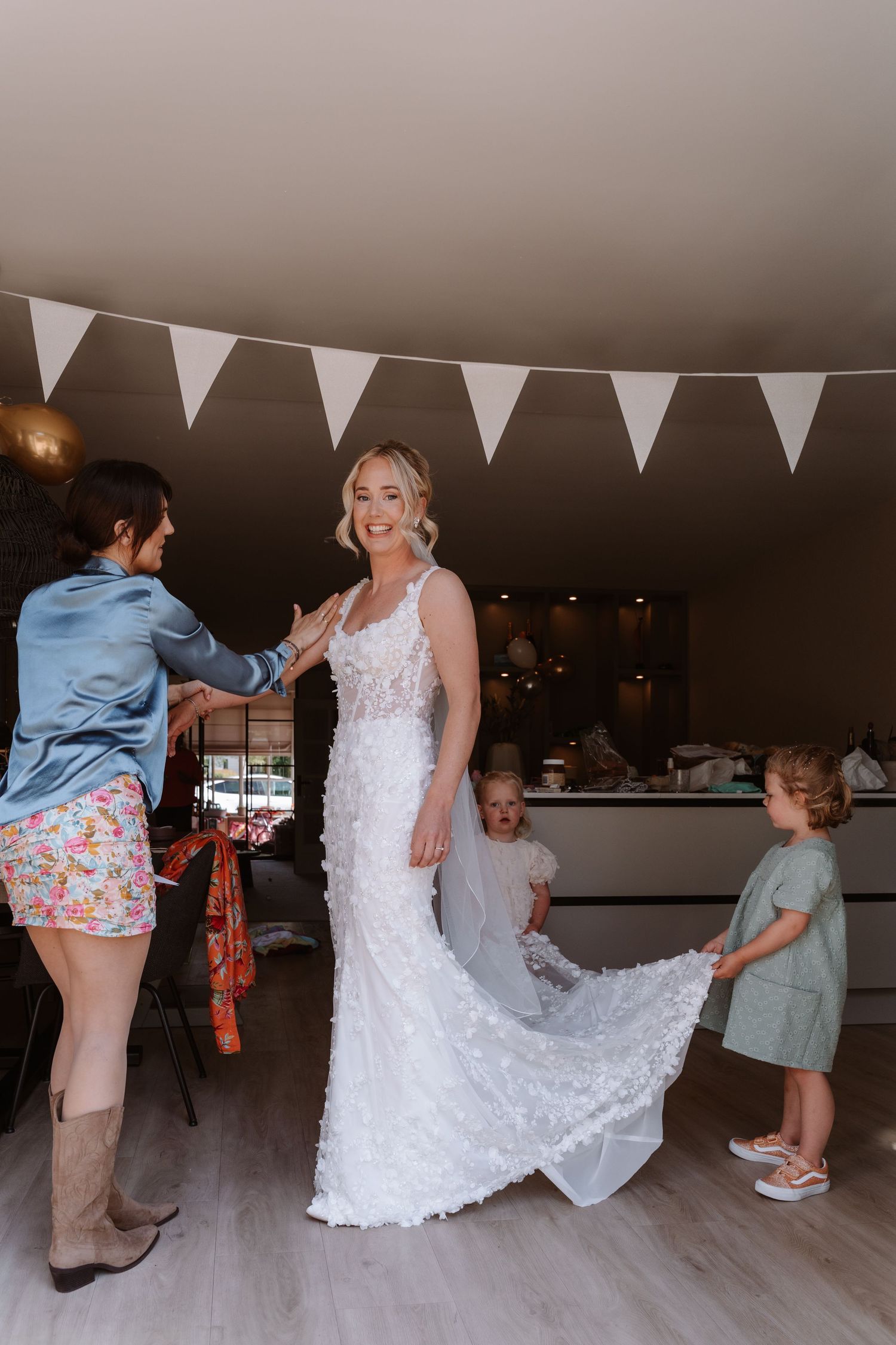 Bride in white lace gown being helped by attendants, including children, in a decorated venue with bunting.