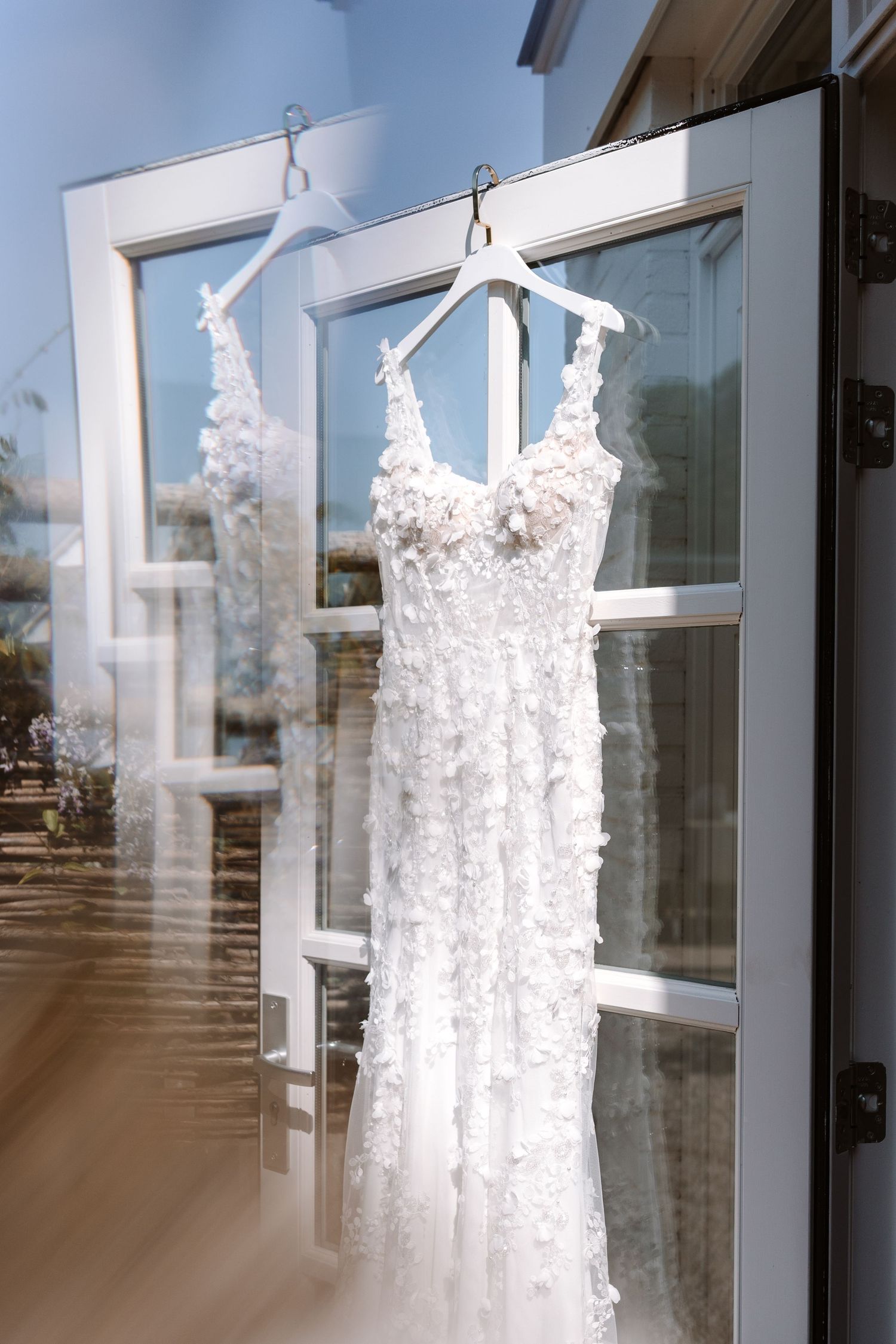 White floral lace wedding dress hanging on a white hanger in front of a bright glass door.