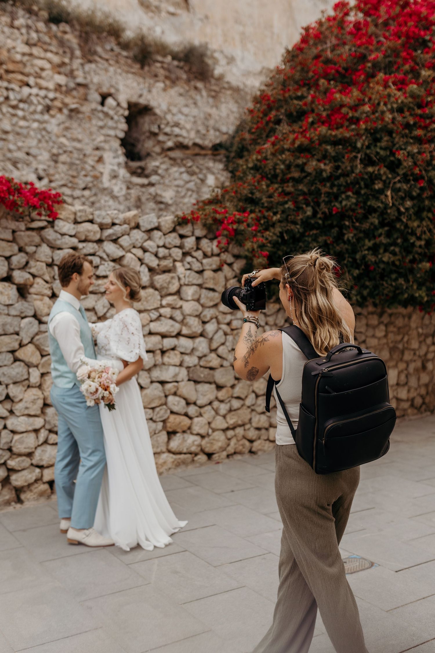 Behind the scenes photographer captures a couple in white and blue outfits against a stone wall with red flowers.