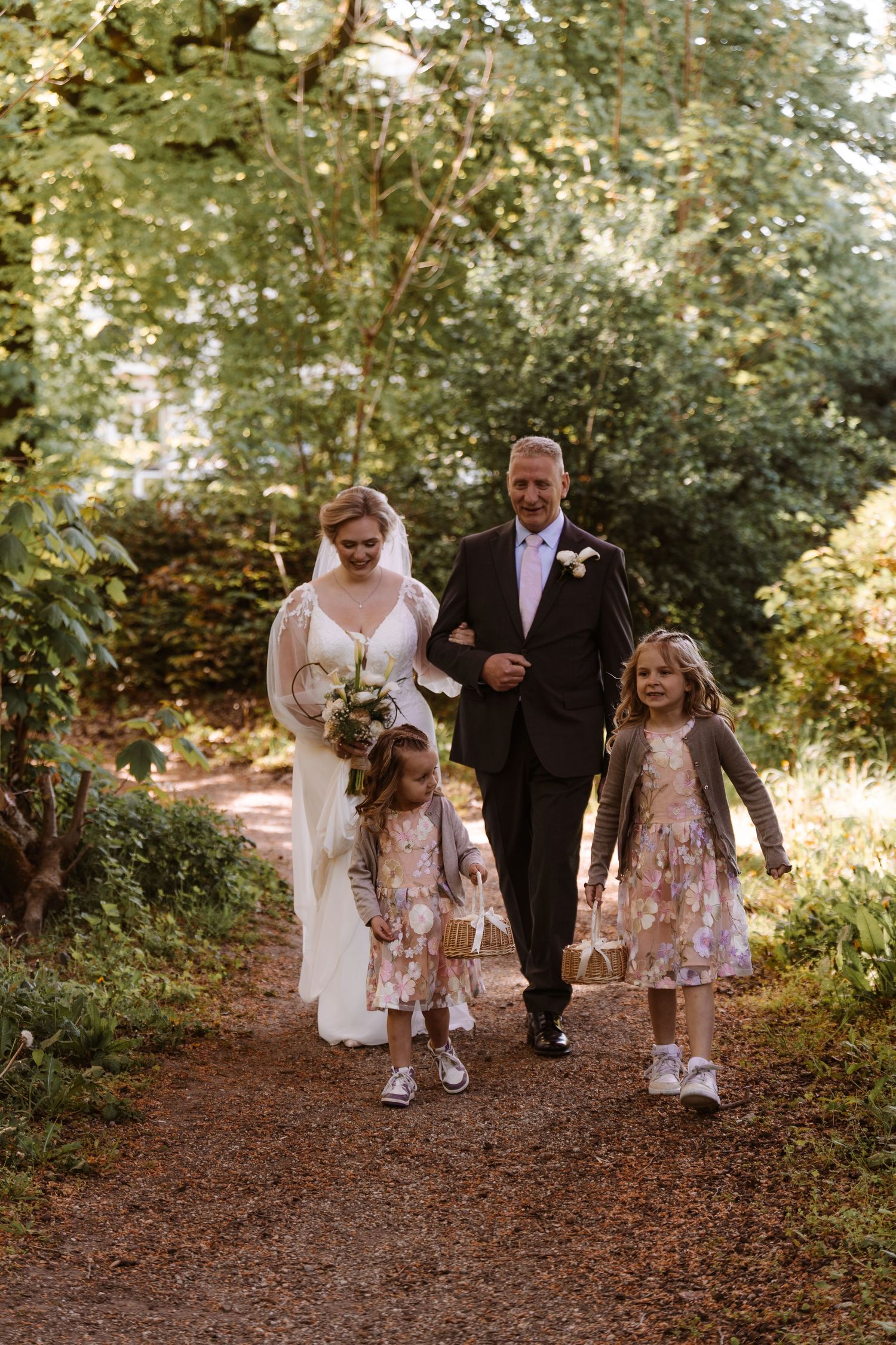 Bride and groom walk with two young flower girls down a tree-lined garden path on their wedding day.