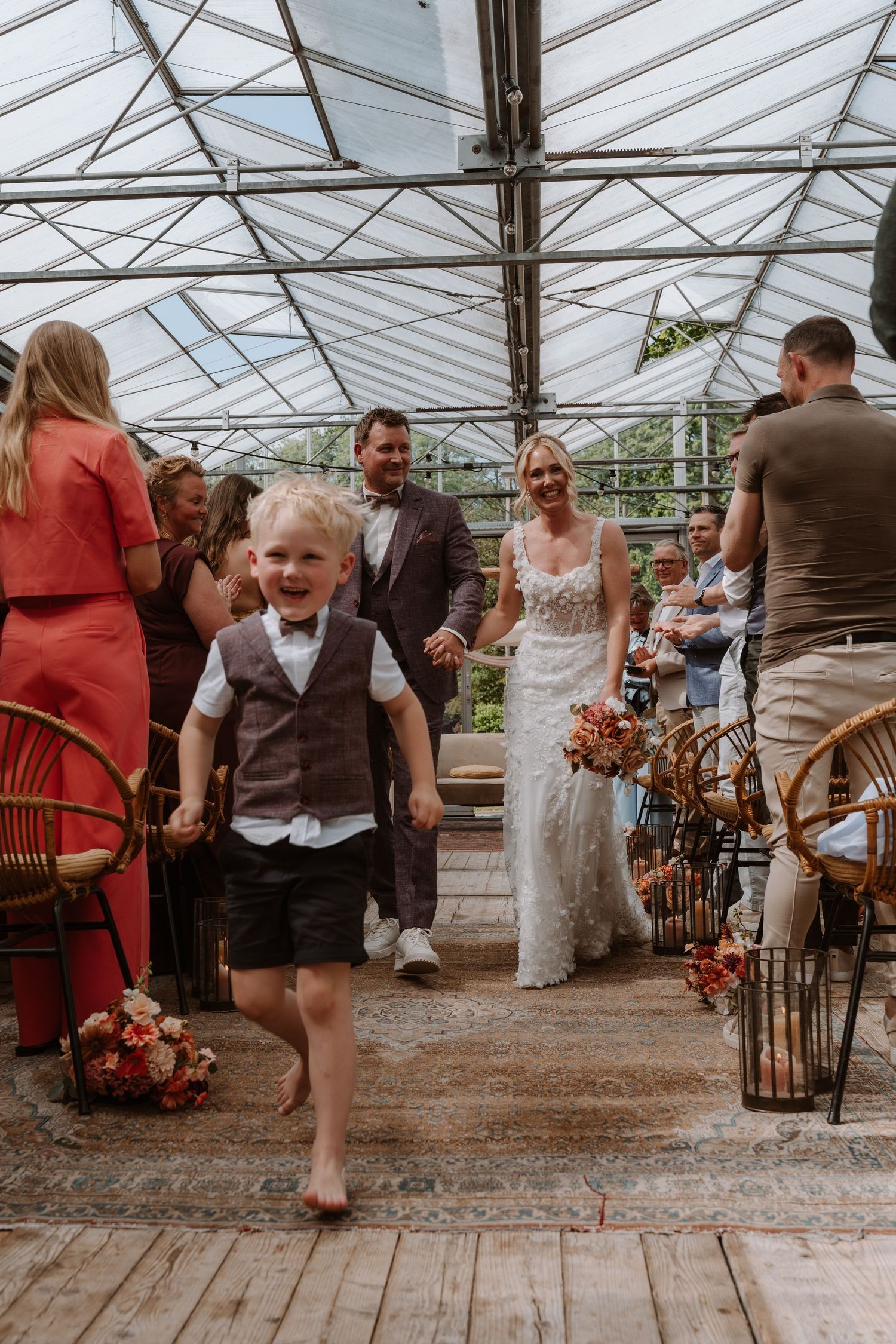 A joyful boy runs ahead of a smiling bride and groom during their wedding ceremony in a greenhouse venue.