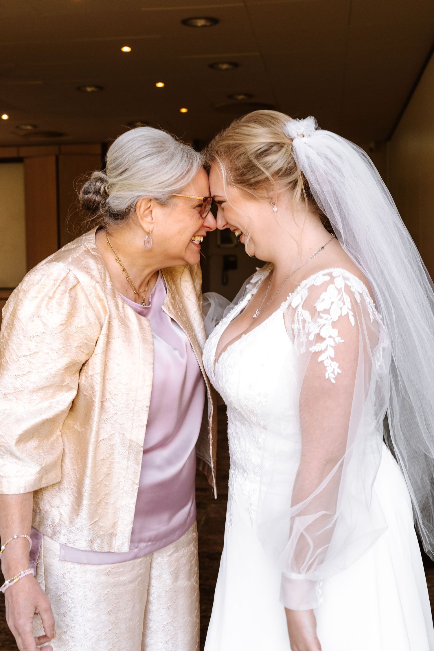 A bride in a white gown and veil shares a tender forehead-to-forehead moment with an older woman in a champagne jacket.
