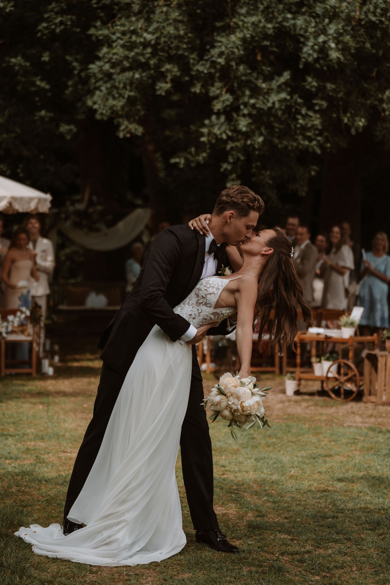 Romantic outdoor wedding reception scene with couple dancing on grass surrounded by guests.