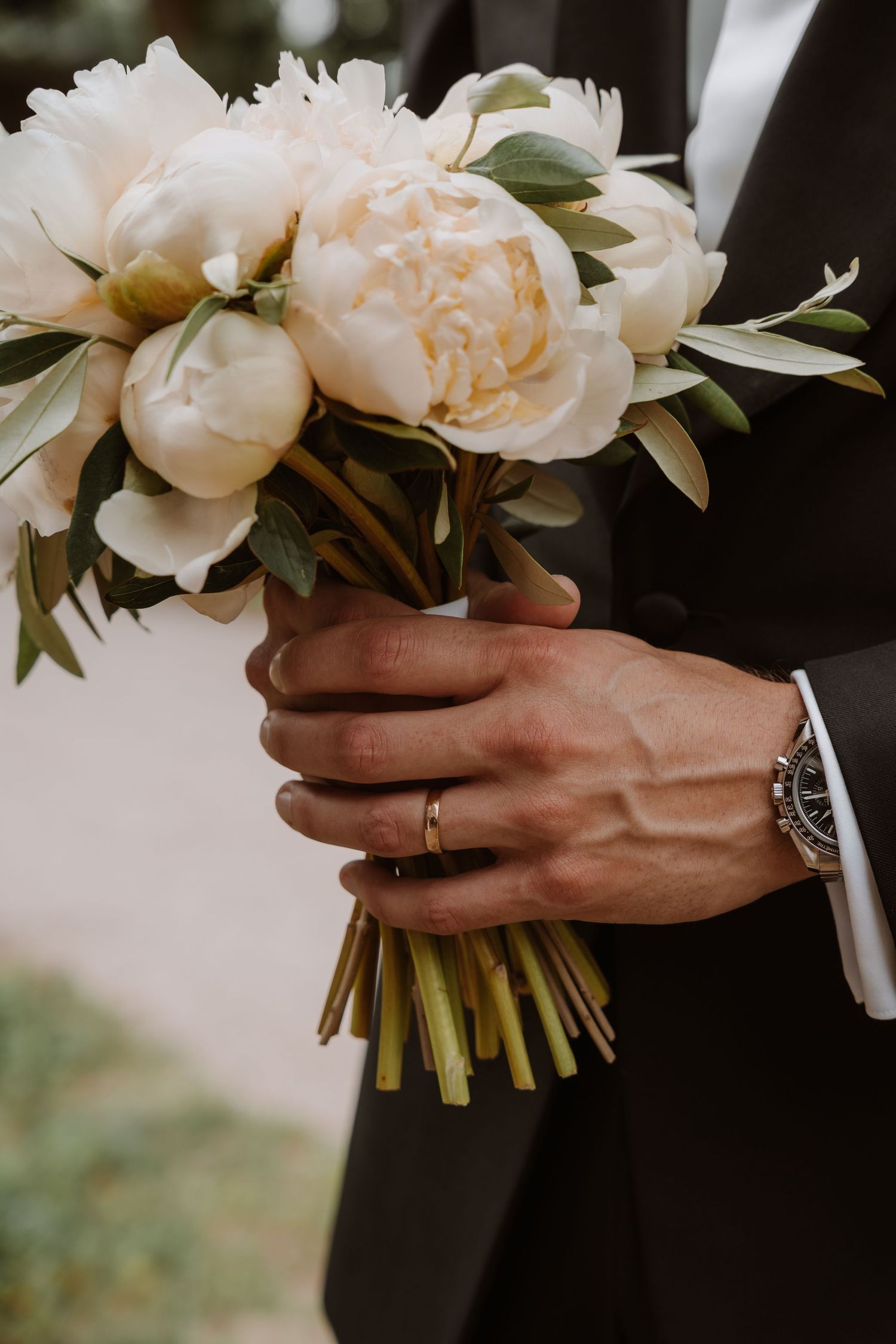 Elegant white peony wedding bouquet held in formal-dressed hands with wedding ring visible.