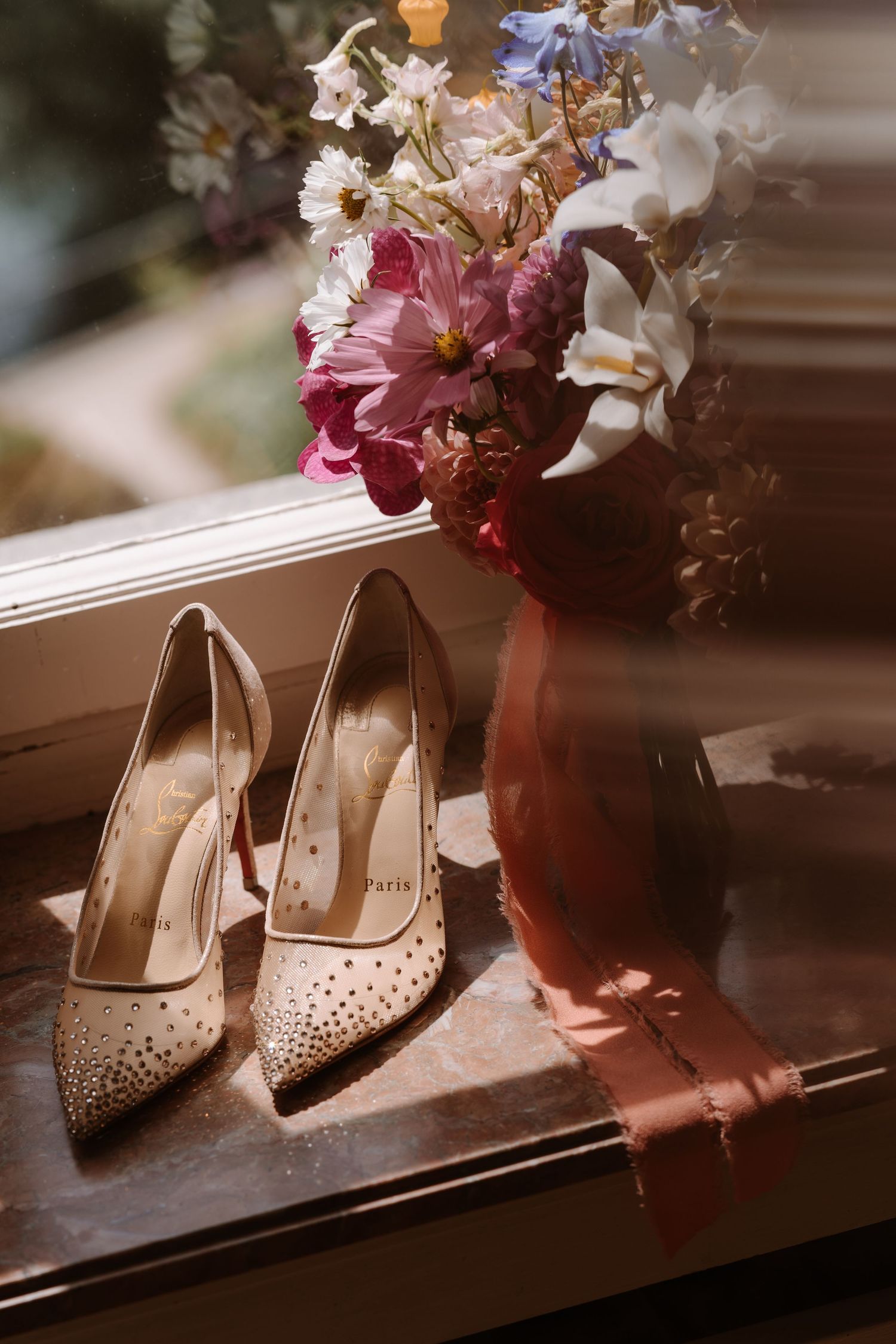 Elegant rhinestone-studded beige heels sit beside a pink floral arrangement in a clay vase on a wooden surface.