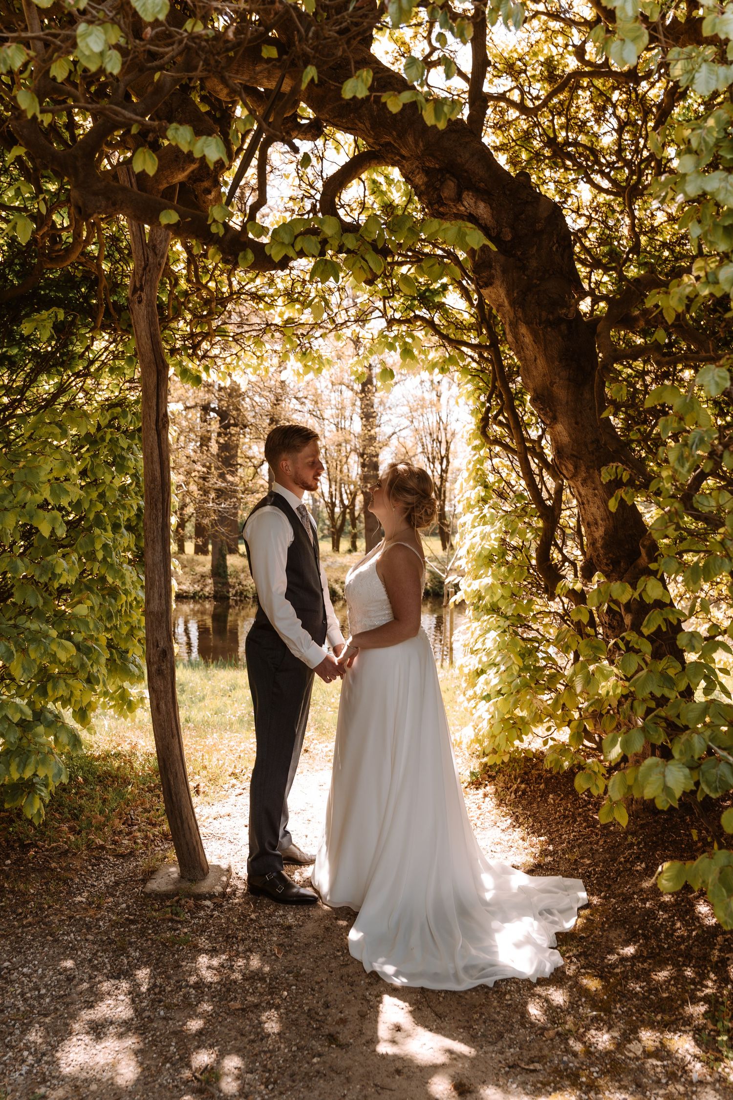 Bride and groom holding hands under a natural tree arch in a sunlit forest during their outdoor wedding ceremony.