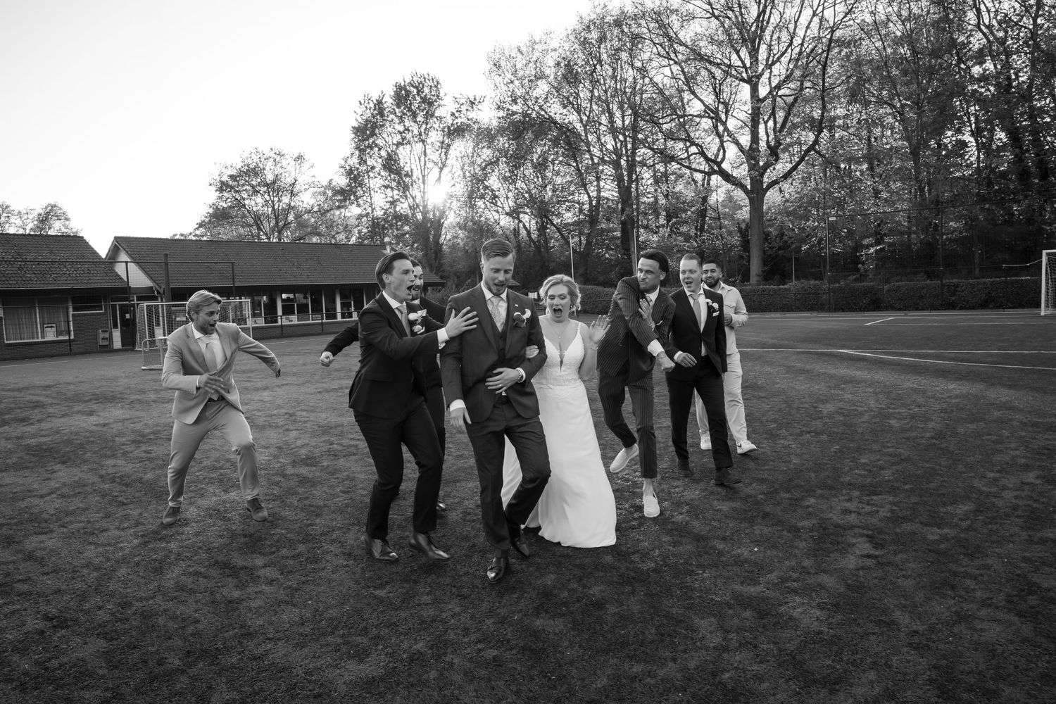Black and white wedding photo of bride, groom, and groomsmen joyfully dancing on a grass field outdoors.