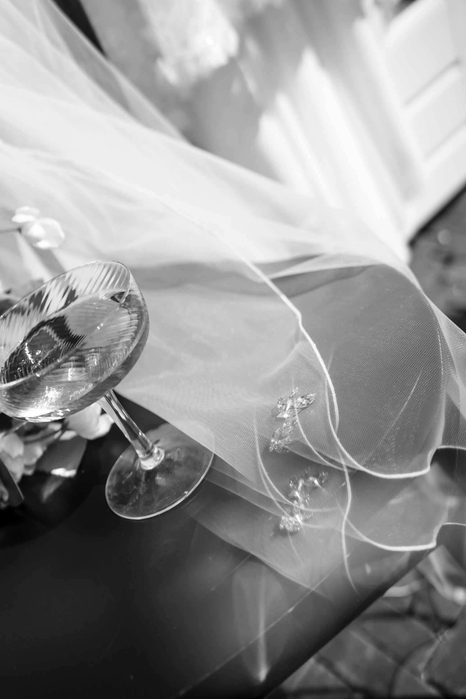 Black and white photo of wedding champagne glasses beside a delicate bridal veil with floral details.